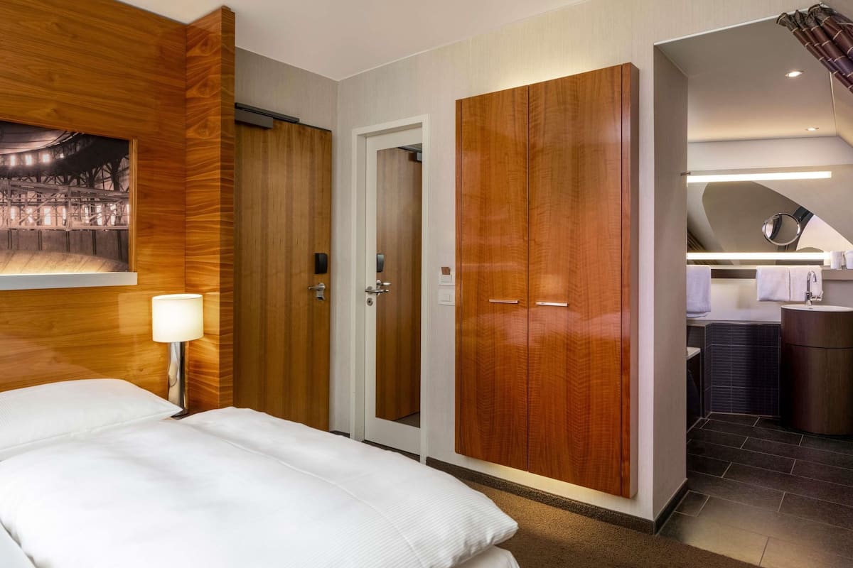 A hotel room with a bed, wooden wardrobe, a bathroom with a mirror, and a painting on the wall.
