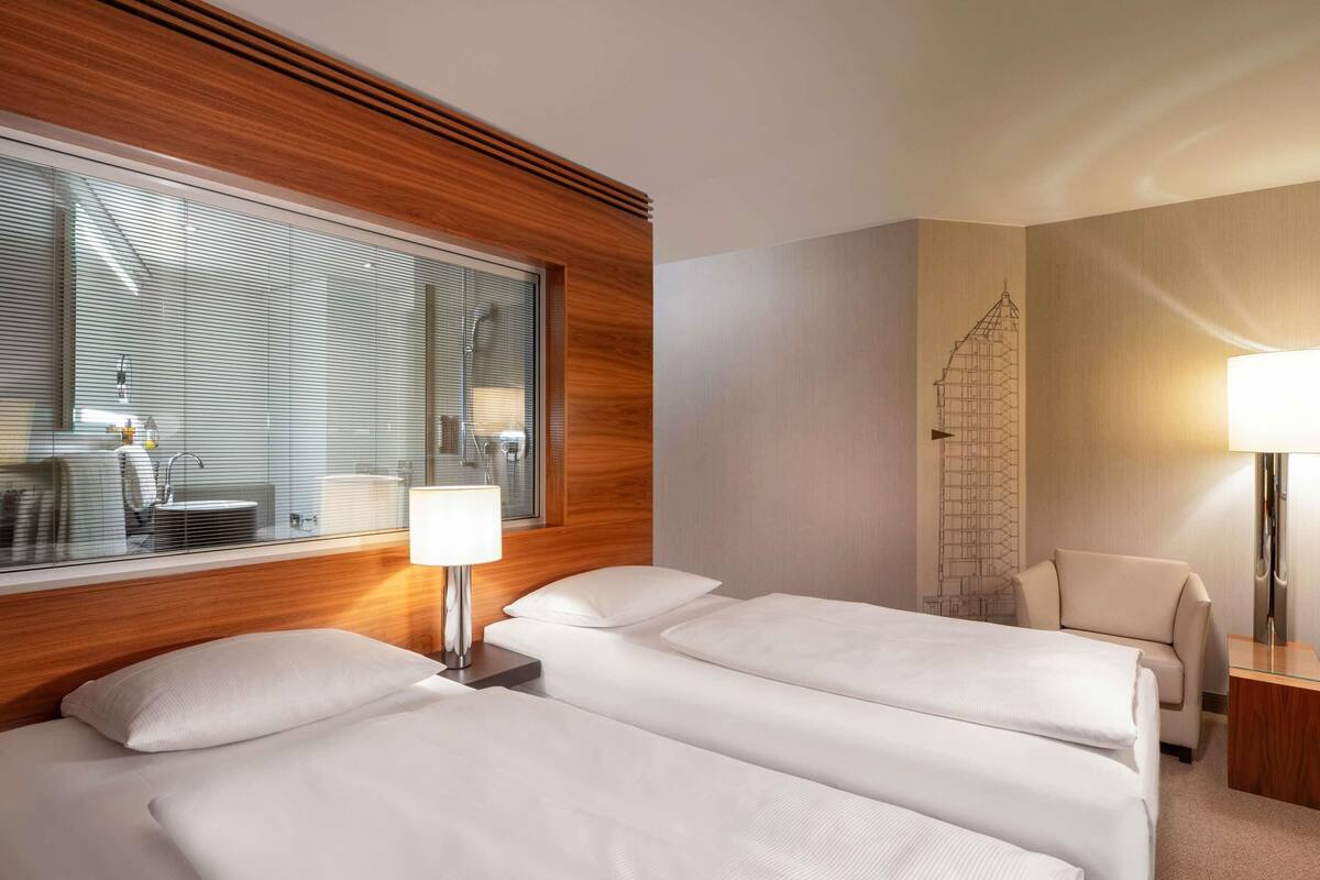 A hotel room with two beds, a wooden headboard, a large window with blinds, a desk, and a chair.