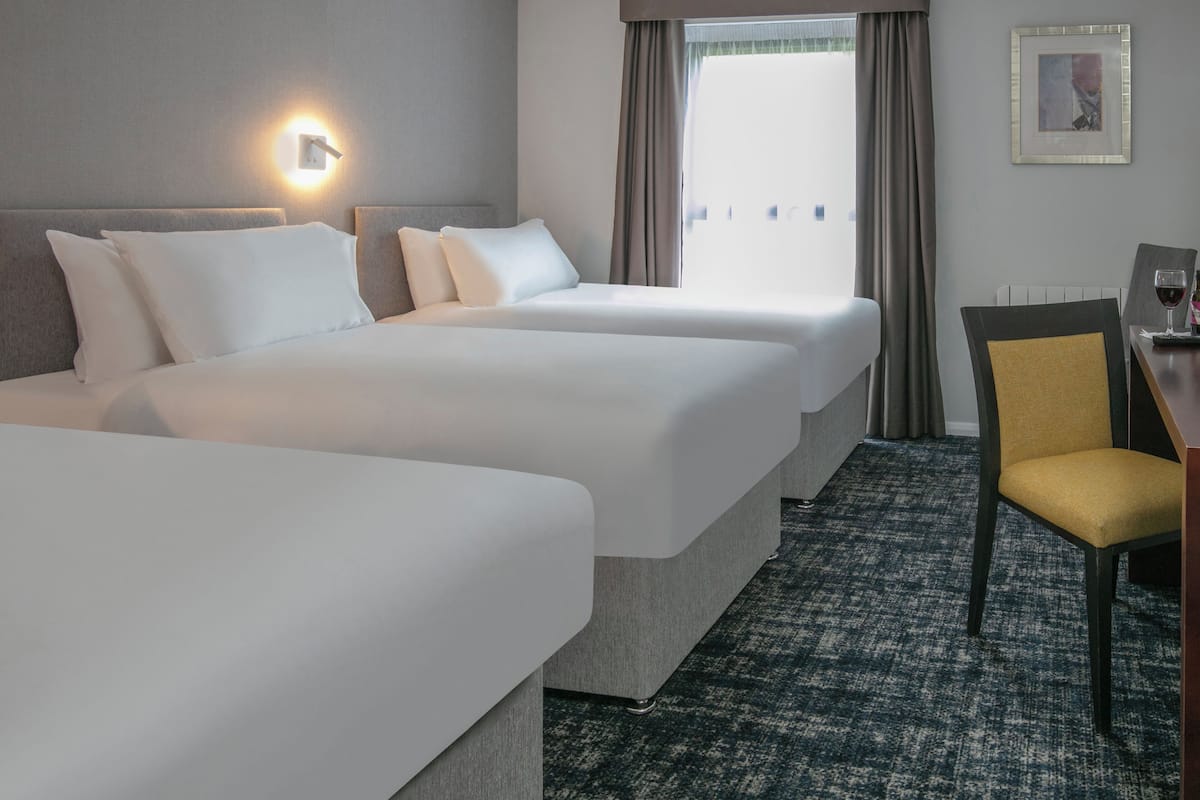 Habitación de hotel con dos camas, un escritorio con silla, una lámpara de pared y un ventanal con cortinas.