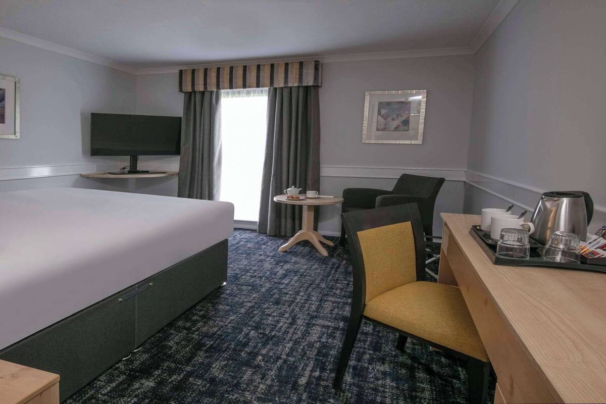 Una habitación de hotel con cama, escritorio con silla, televisión, mesa de centro y ventana con cortinas.
