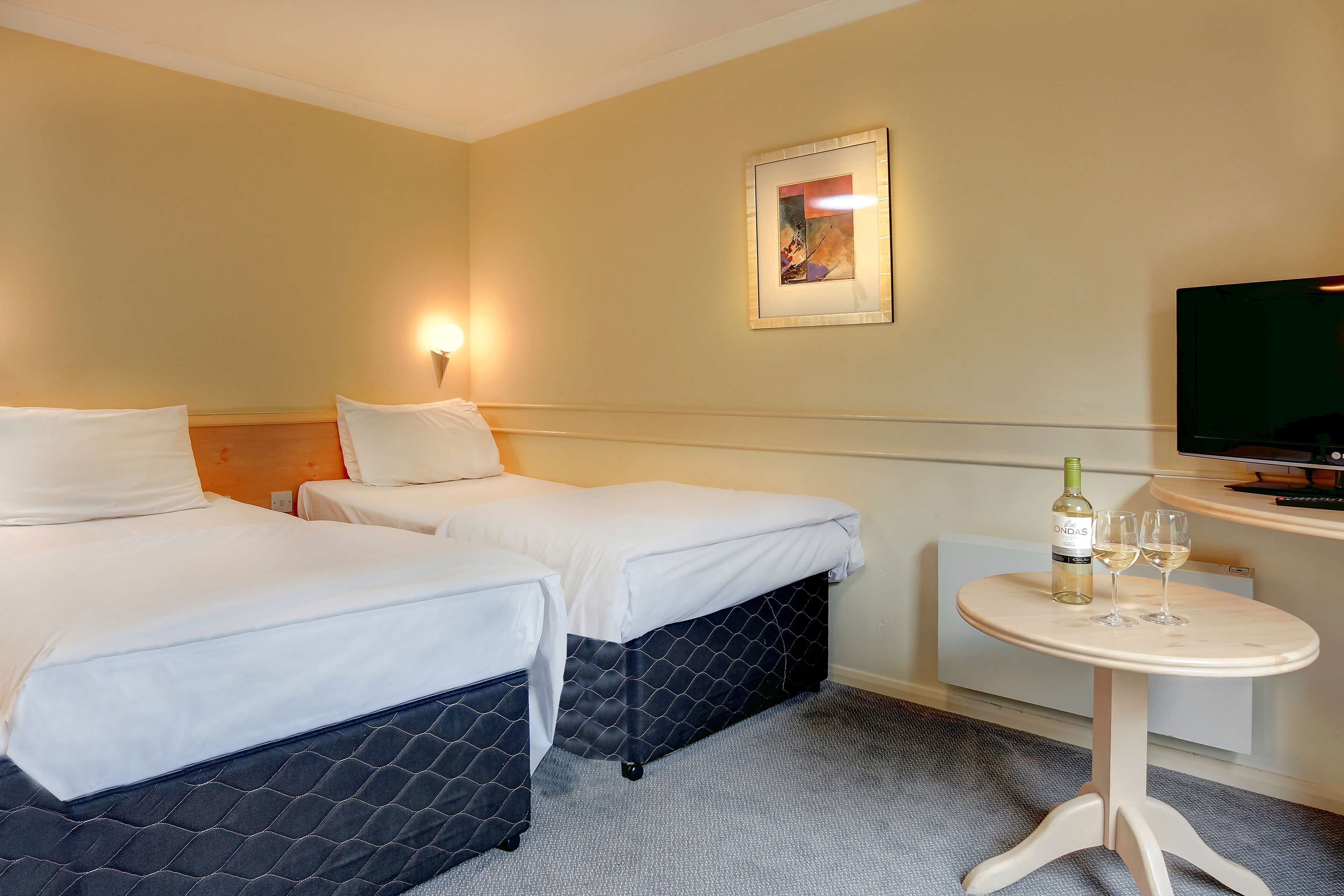 Une chambre d’hôtel avec deux lits, une petite table sur laquelle se trouvent une bouteille et des verres, une télévision et un tableau encadré au mur.