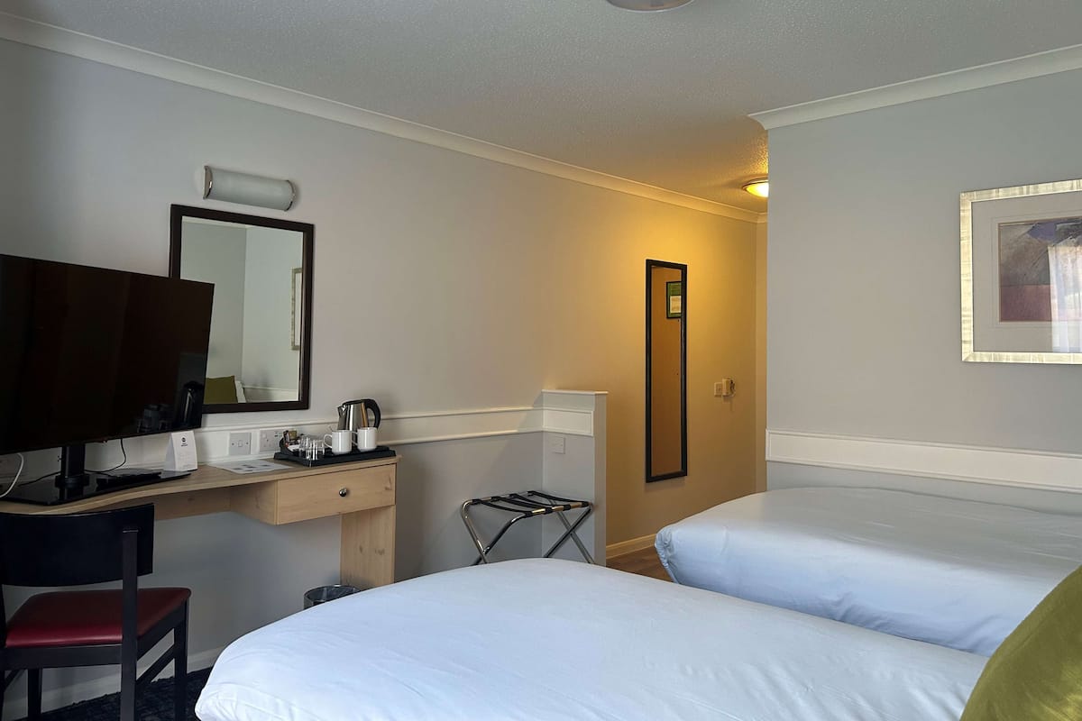 Habitación de hotel con dos camas, un escritorio con ordenador, una silla, un espejo, una plancha pequeña y un cuadro en la pared.