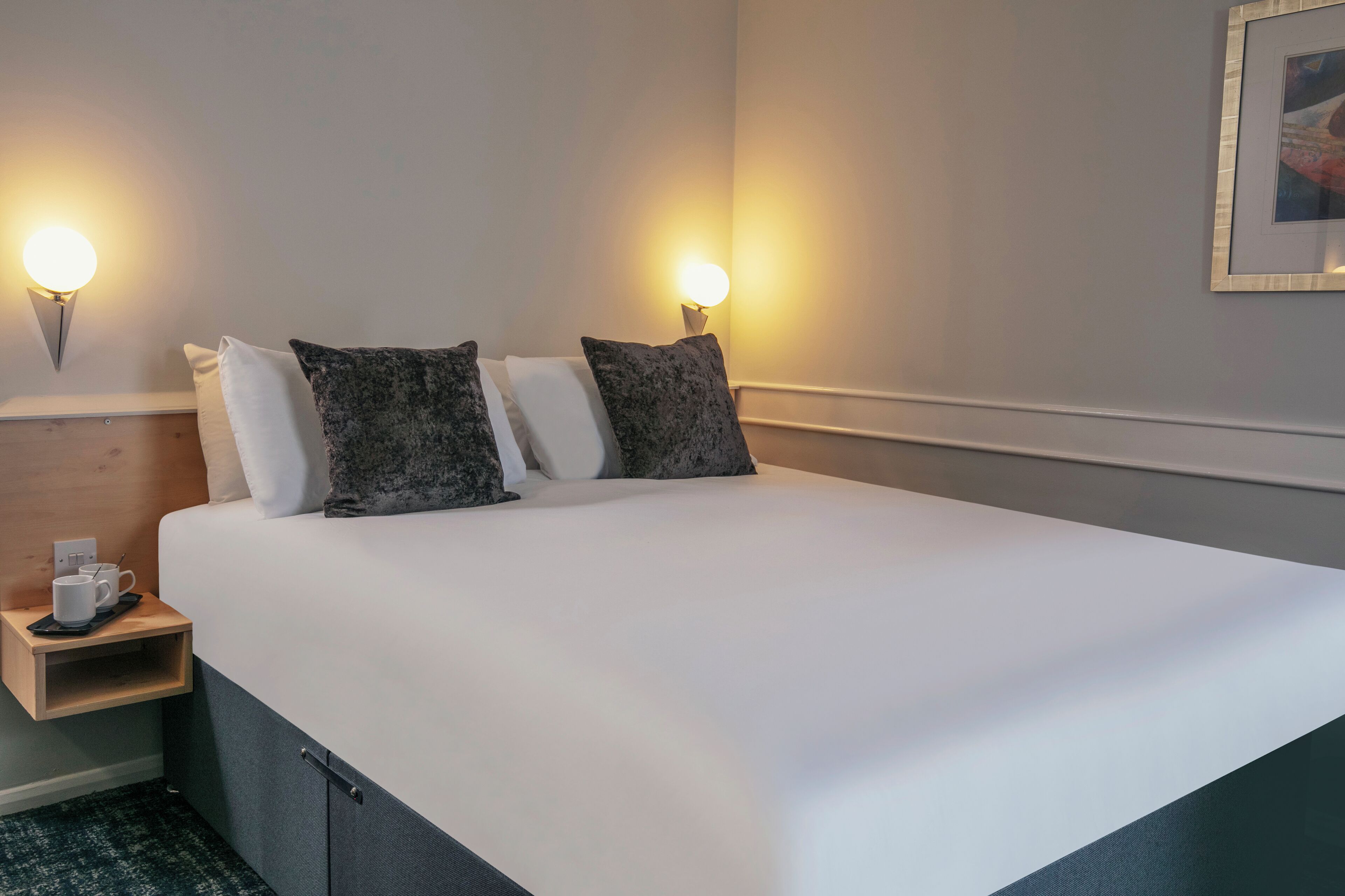 A neatly made bed with white and dark grey pillows, a small bedside table with a cup and saucer, and wall-mounted lamps.