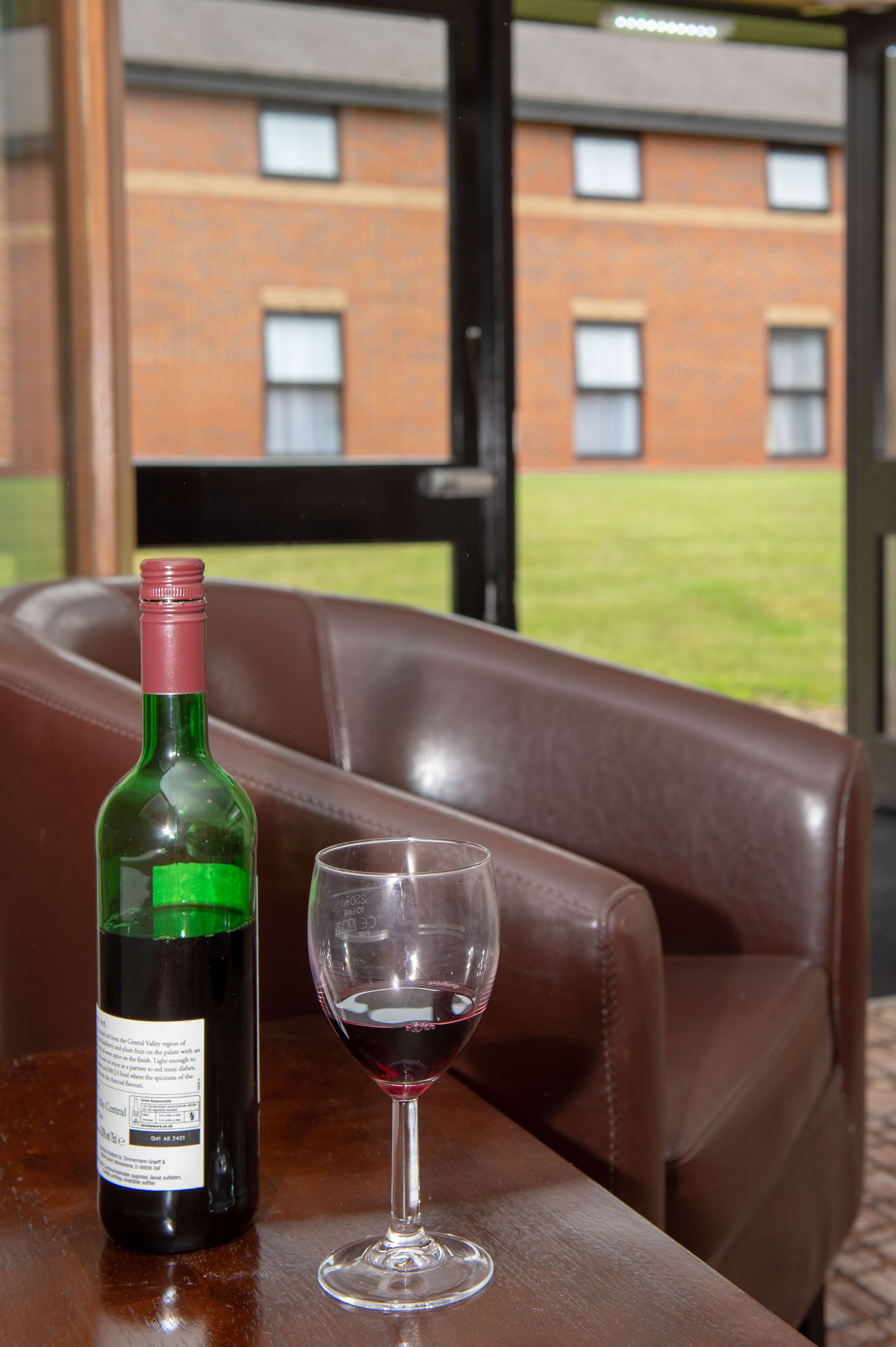 A bottle of wine and an empty glass on a table in a room with a leather couch and a view of a brick building through a window.