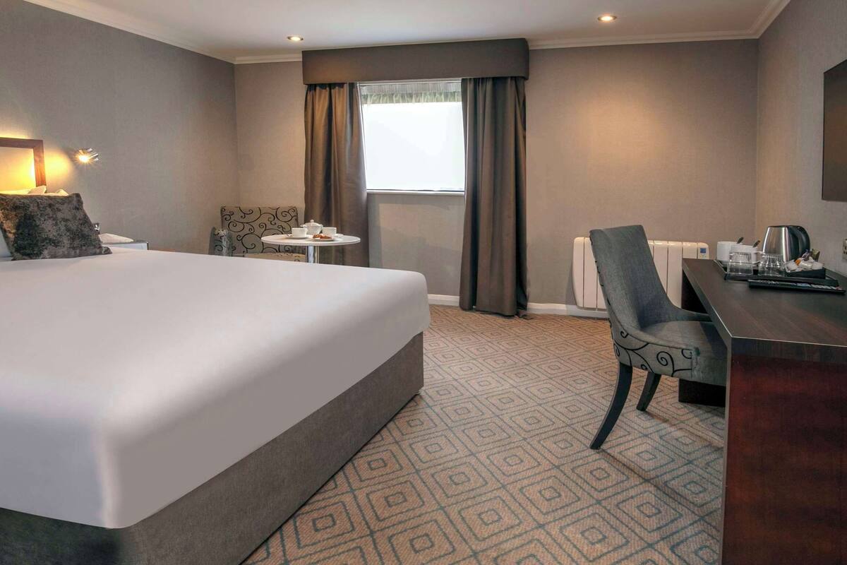 Habitación de hotel con una cama grande, un escritorio, una silla, una mesita con una taza y un televisor en la pared.