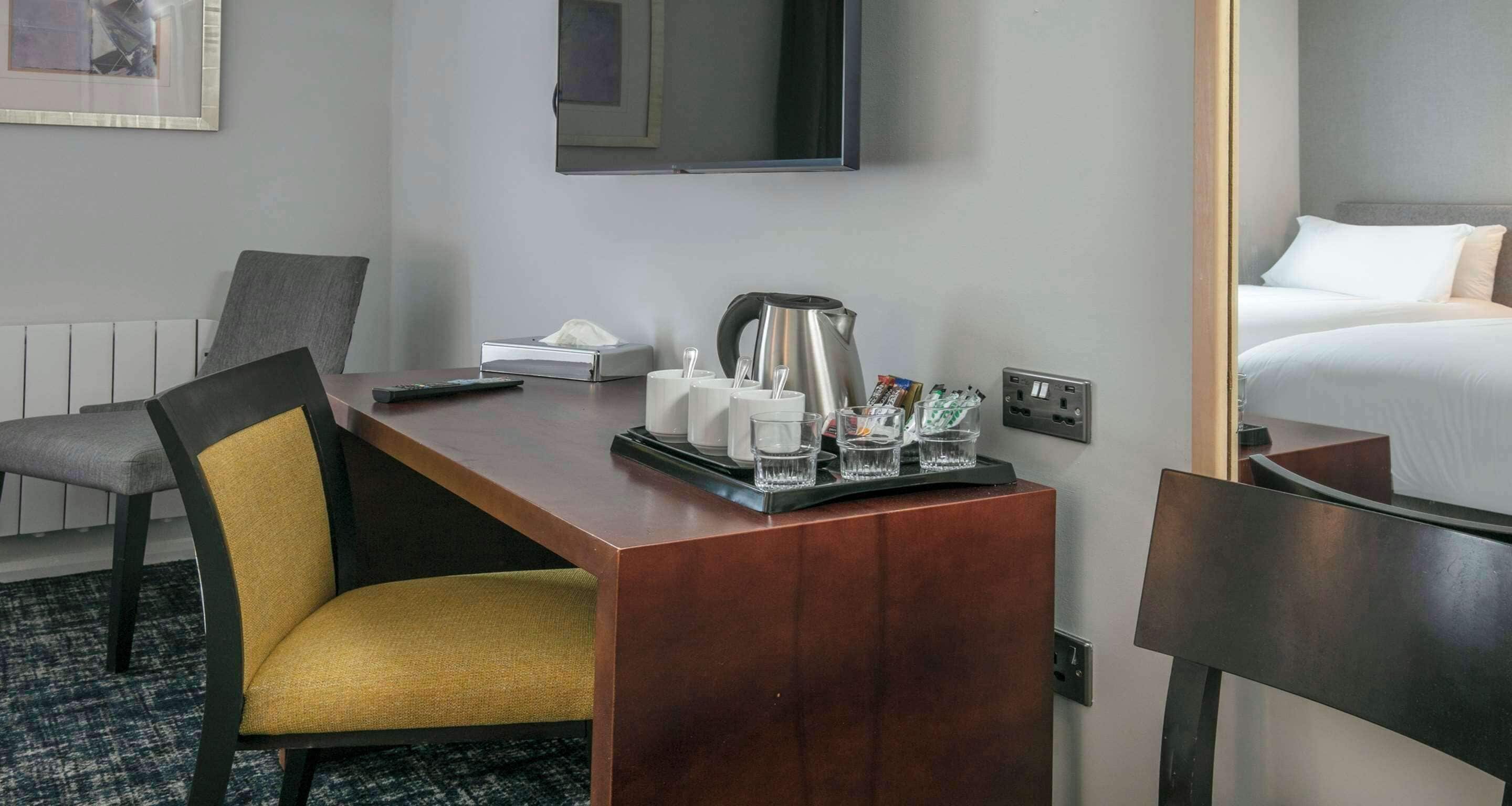 A hotel room with a wooden desk, a chair, a bed, a TV mounted on the wall, and a small table with a coffee maker and cups.