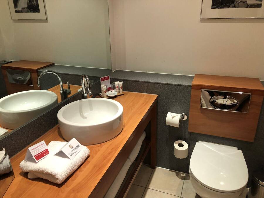 A modern bathroom with a double sink, a mirror, a toilet, and a small shelf with toiletries.