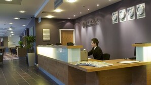 A reception desk with a wooden counter, a laptop, and some documents. There are clocks on the wall and a sign above the desk. - Ramada Plaza by WyndhamWrexham (Wrexham)