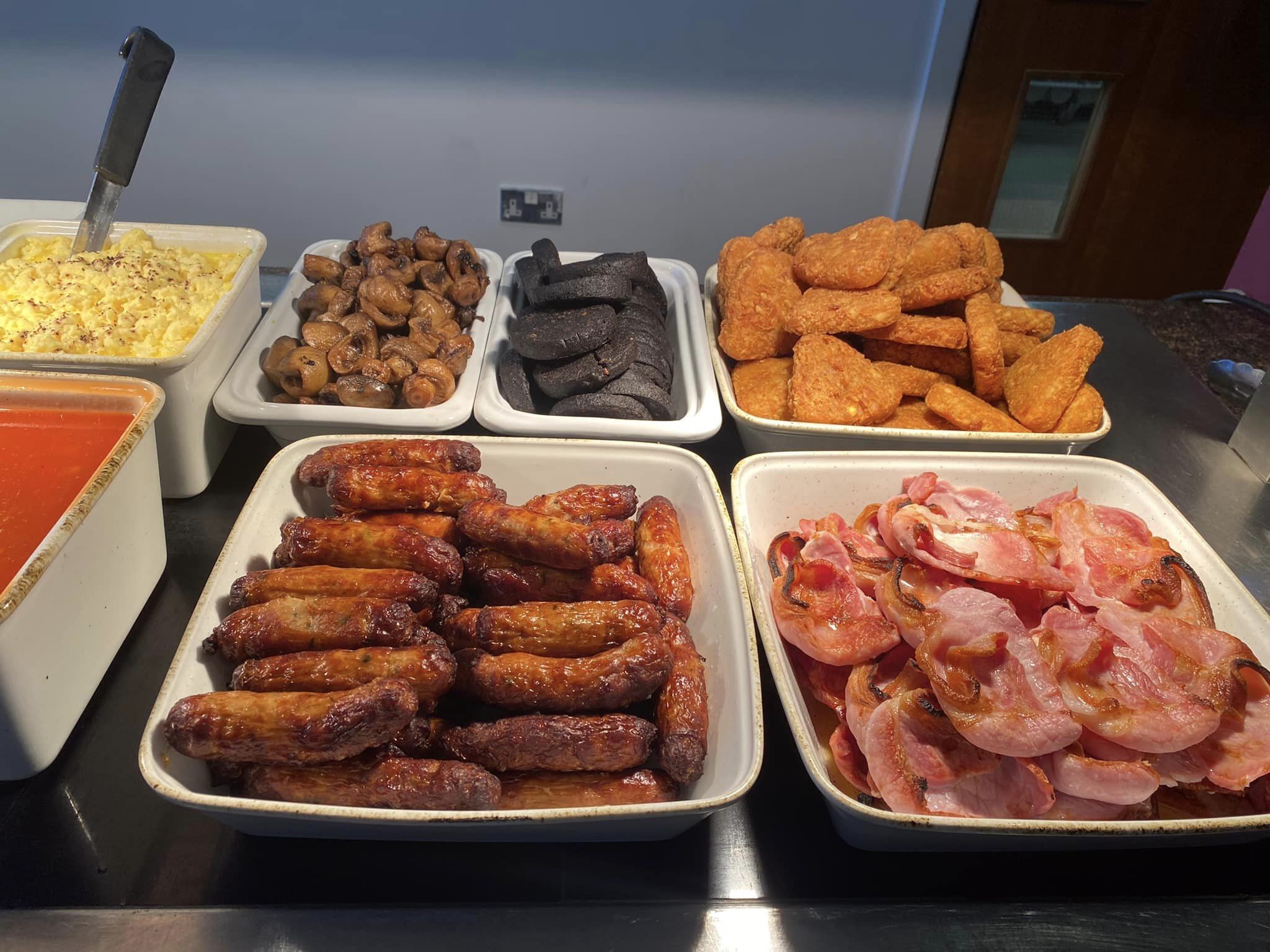 A buffet with various food items including macaroni and cheese, mushrooms, black pudding, fried chicken, sausages, and bacon.