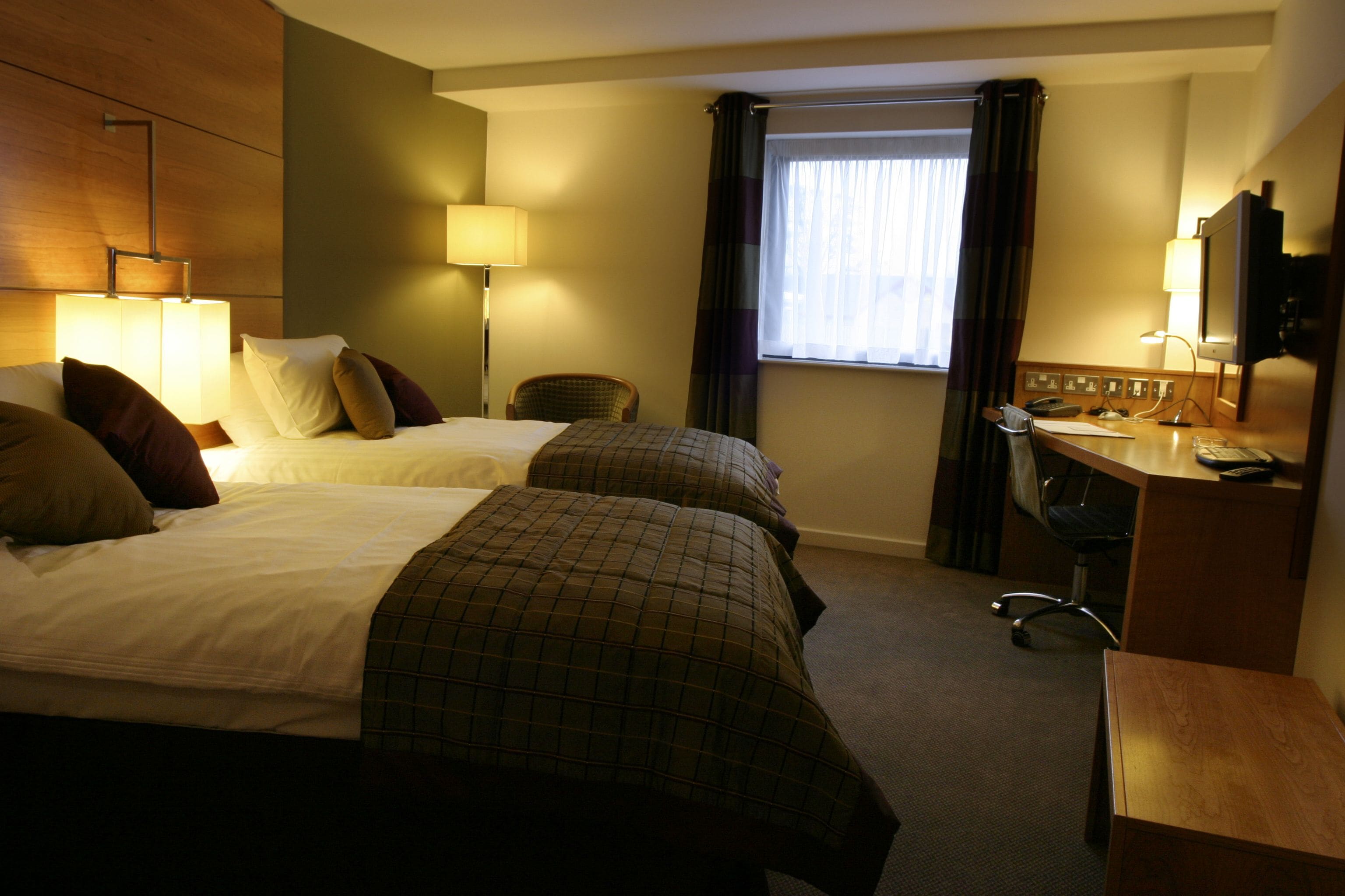 A hotel room with two beds, a desk, a chair, a TV, and a window with curtains.