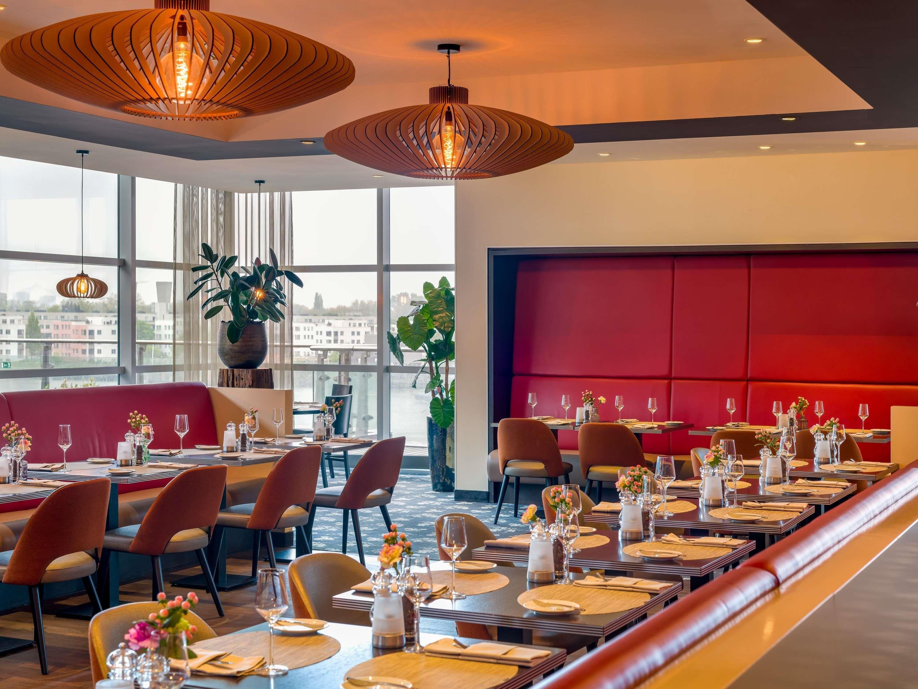 A restaurant with a long dining table set for a meal, large windows offering a view of the city, and modern pendant lighting.