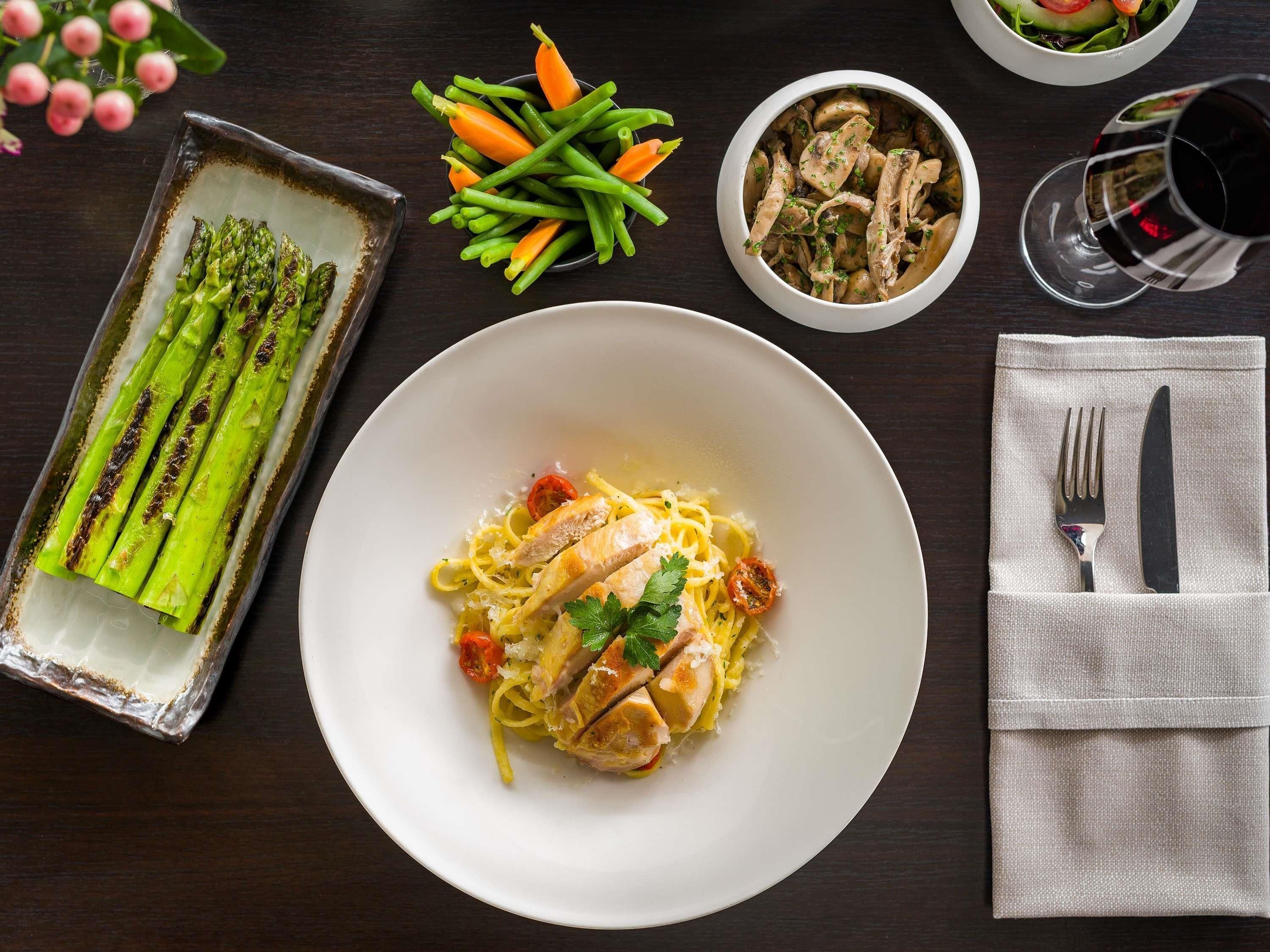 A dining table with a plate of pasta, asparagus, and carrots, a bowl of mushrooms, and a glass of red wine.