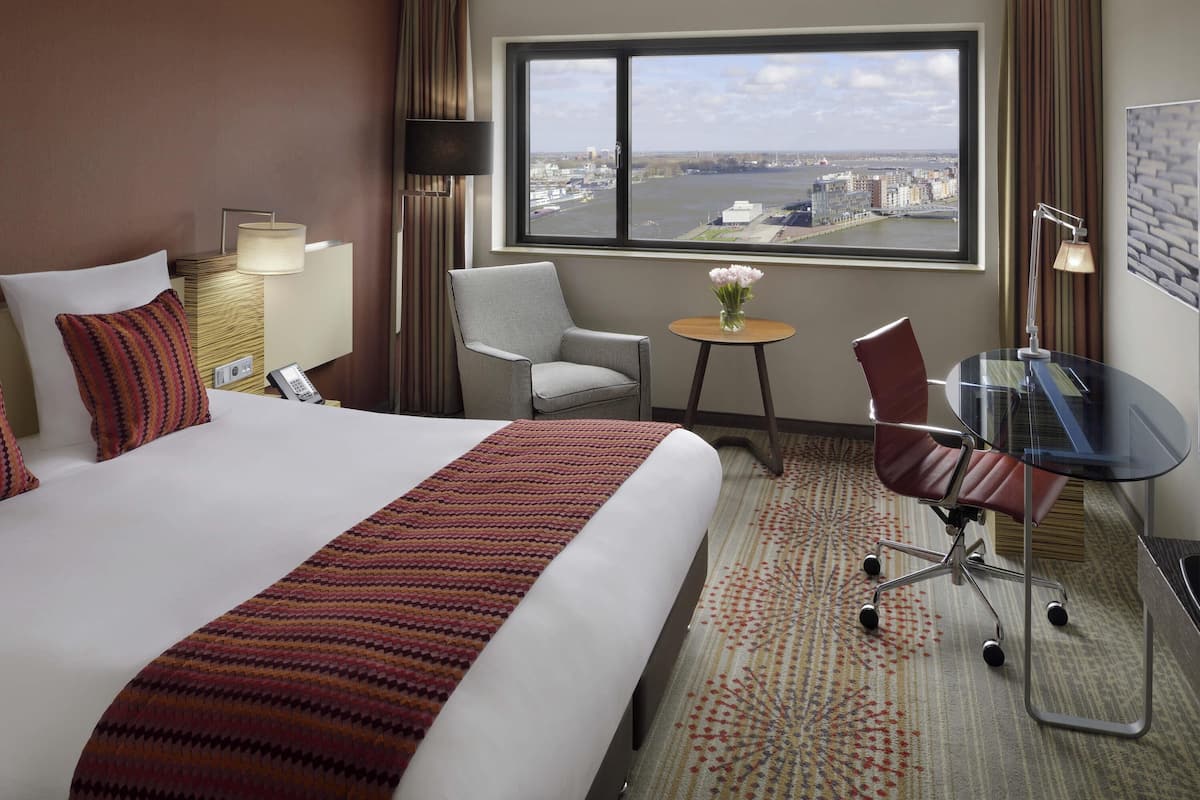 A hotel room with a large bed, a desk, a chair, a small table with flowers, and a view of a cityscape through a large window.