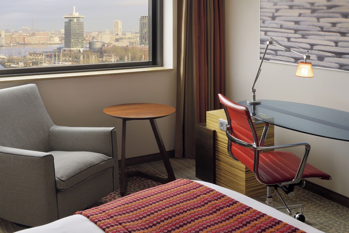 Habitación de hotel con vista a la ciudad, un escritorio, una silla, una cama y una pequeña mesa.