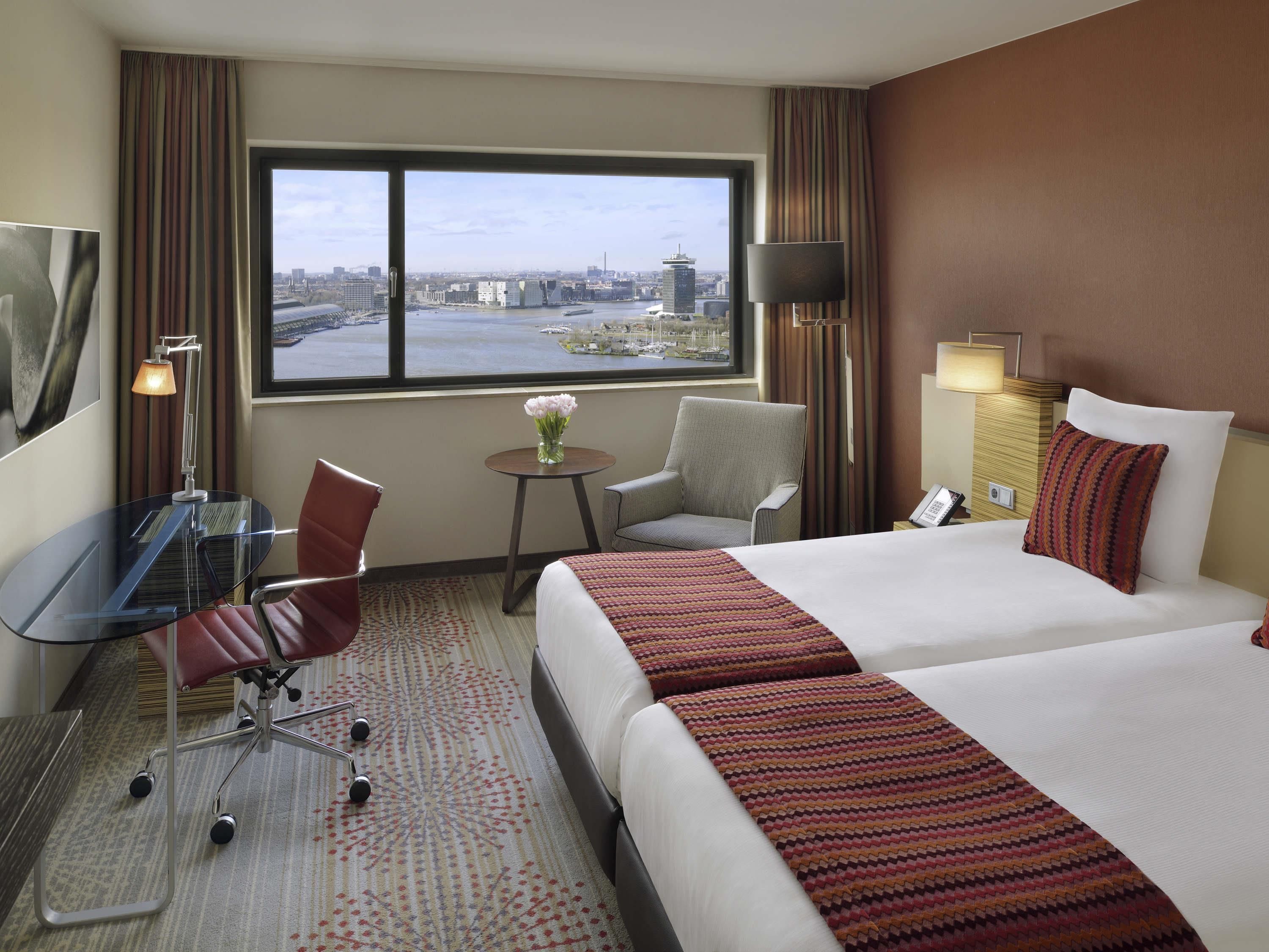 Ein Hotelzimmer mit einem großen Bett, einem Schreibtisch mit Stuhl, einem kleinen Tisch mit Blumenvase und Blick auf eine Stadtlandschaft durch ein großes Fenster.