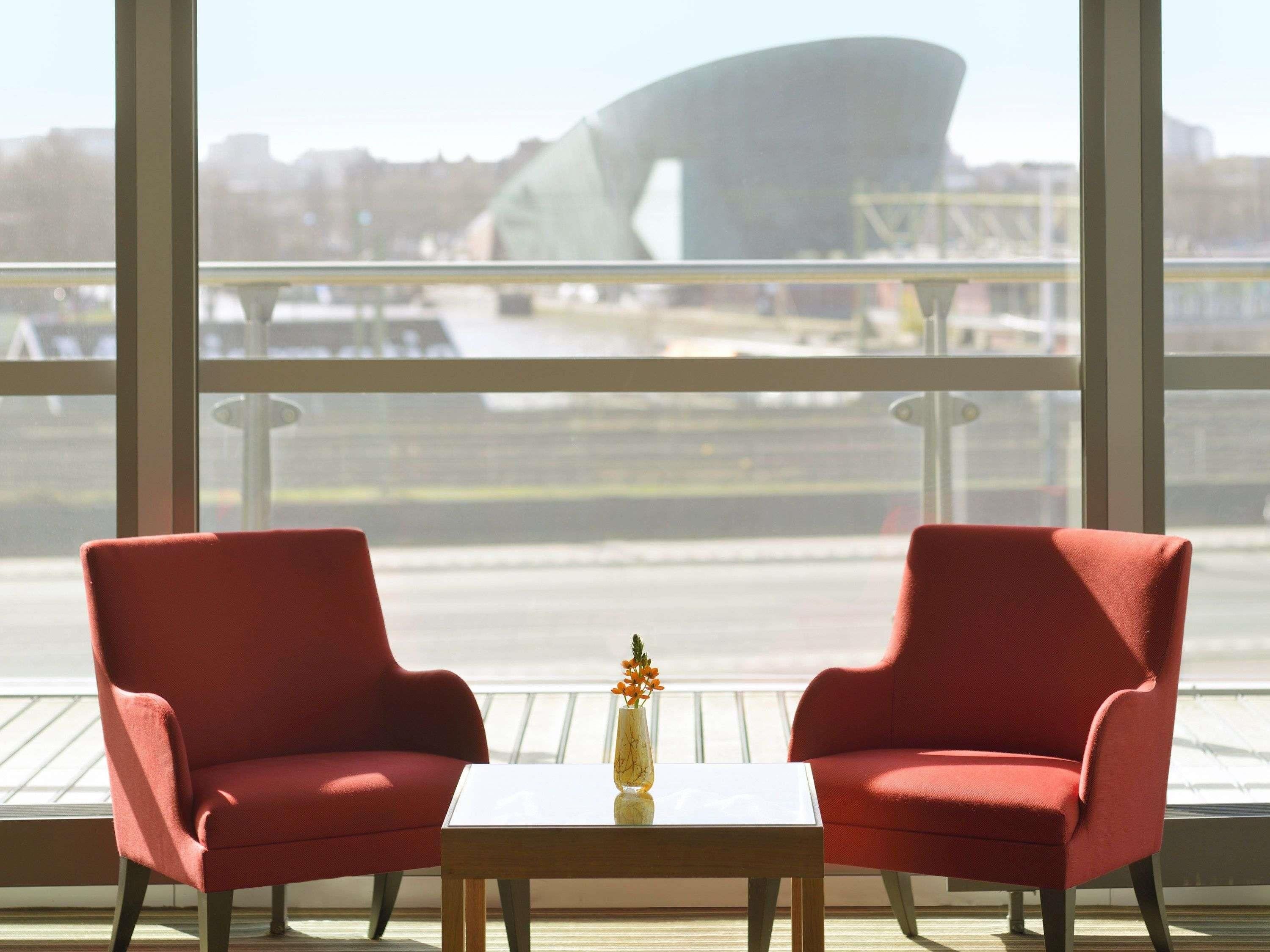 Two red chairs, a small table with a vase, and a large window with a view of a bridge and buildings.