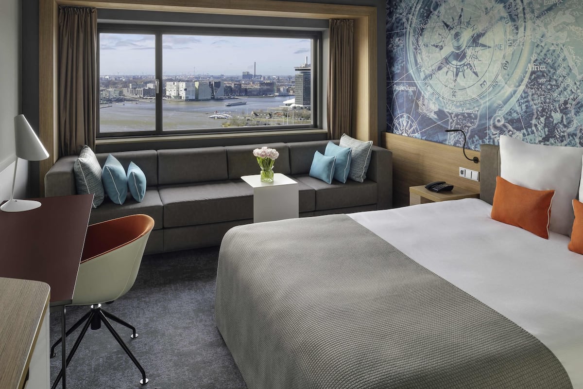 A hotel room with a large bed, a sofa, a desk, and a view of a cityscape through a large window.