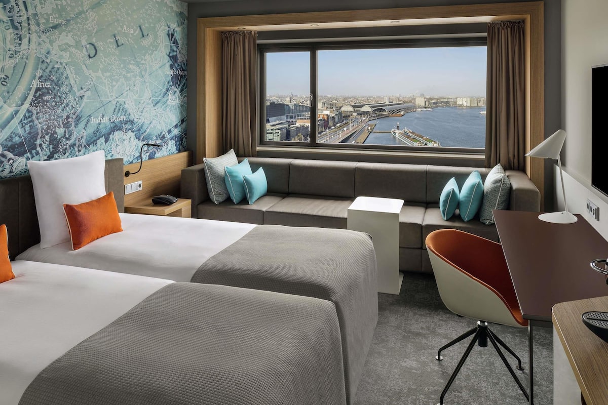 A hotel room with a bed, a desk, a chair, a sofa, and a large window with a city view.