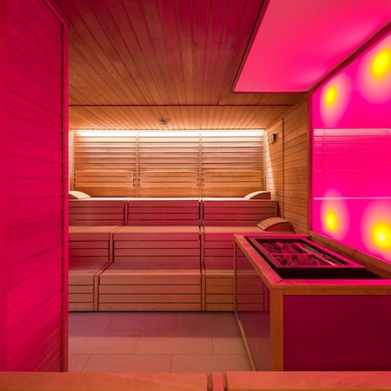 A sauna with wooden benches and a heated floor, surrounded by wooden walls and a pink-lit wall.