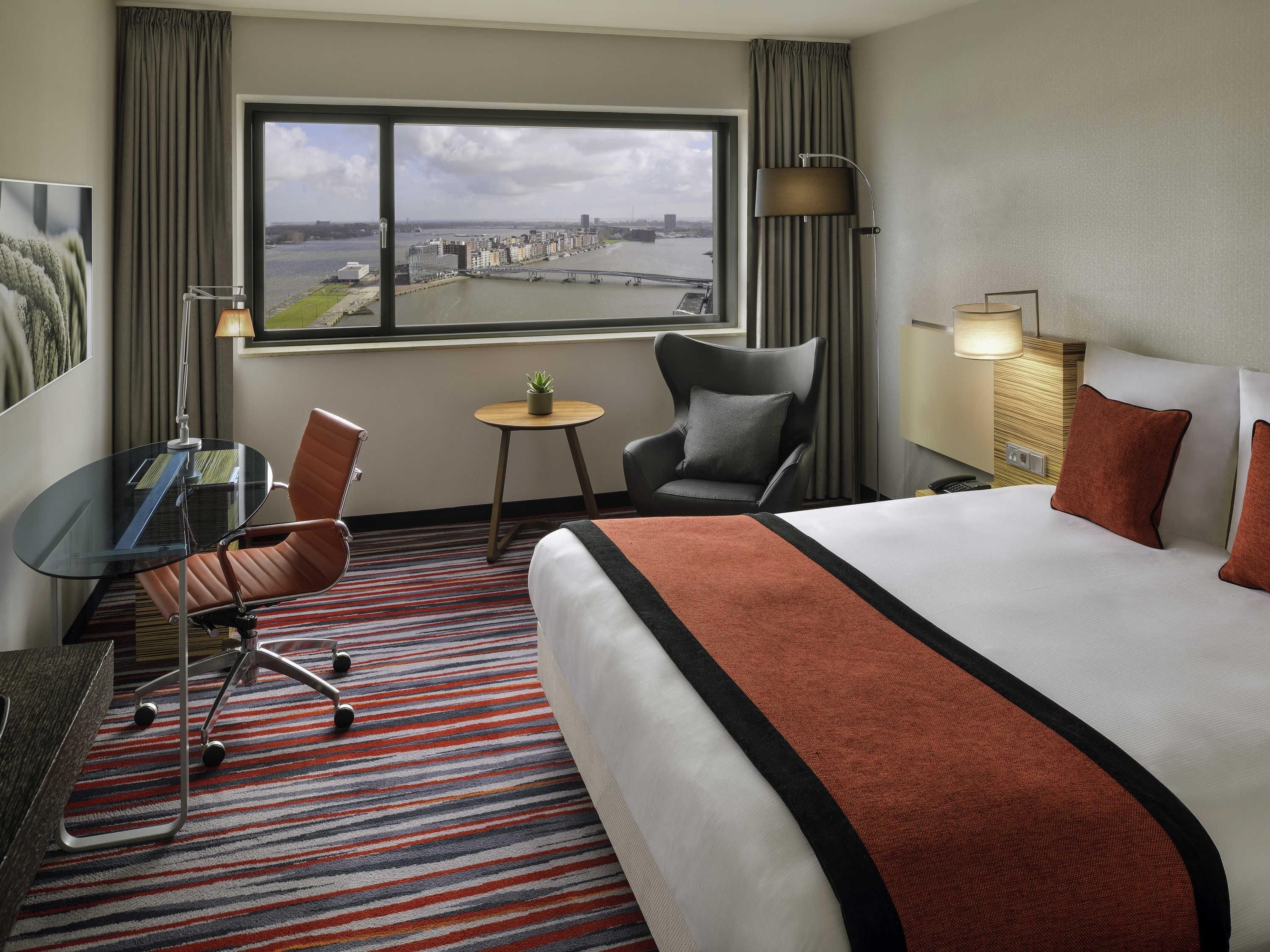 A hotel room with a large bed, a desk, a chair, a small table with a plant, and a view of a cityscape through a window.