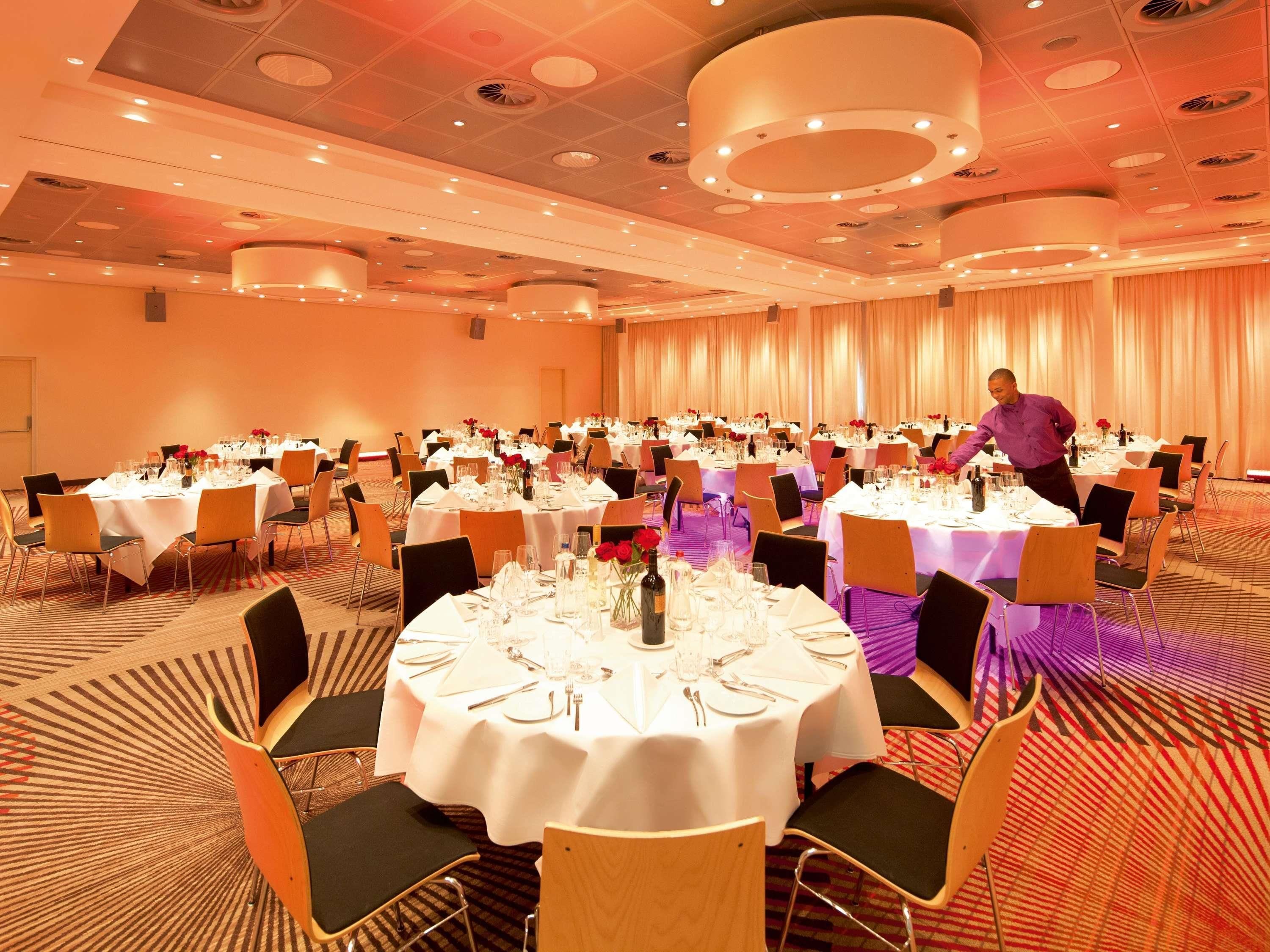 A banquet hall with round tables set for dining, a waiter attending to guests, and a carpeted floor.