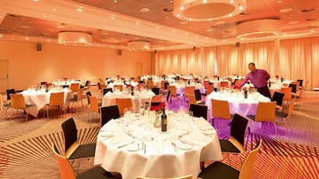 A banquet hall with round tables set for dining, a waiter attending to guests, and a carpeted floor.