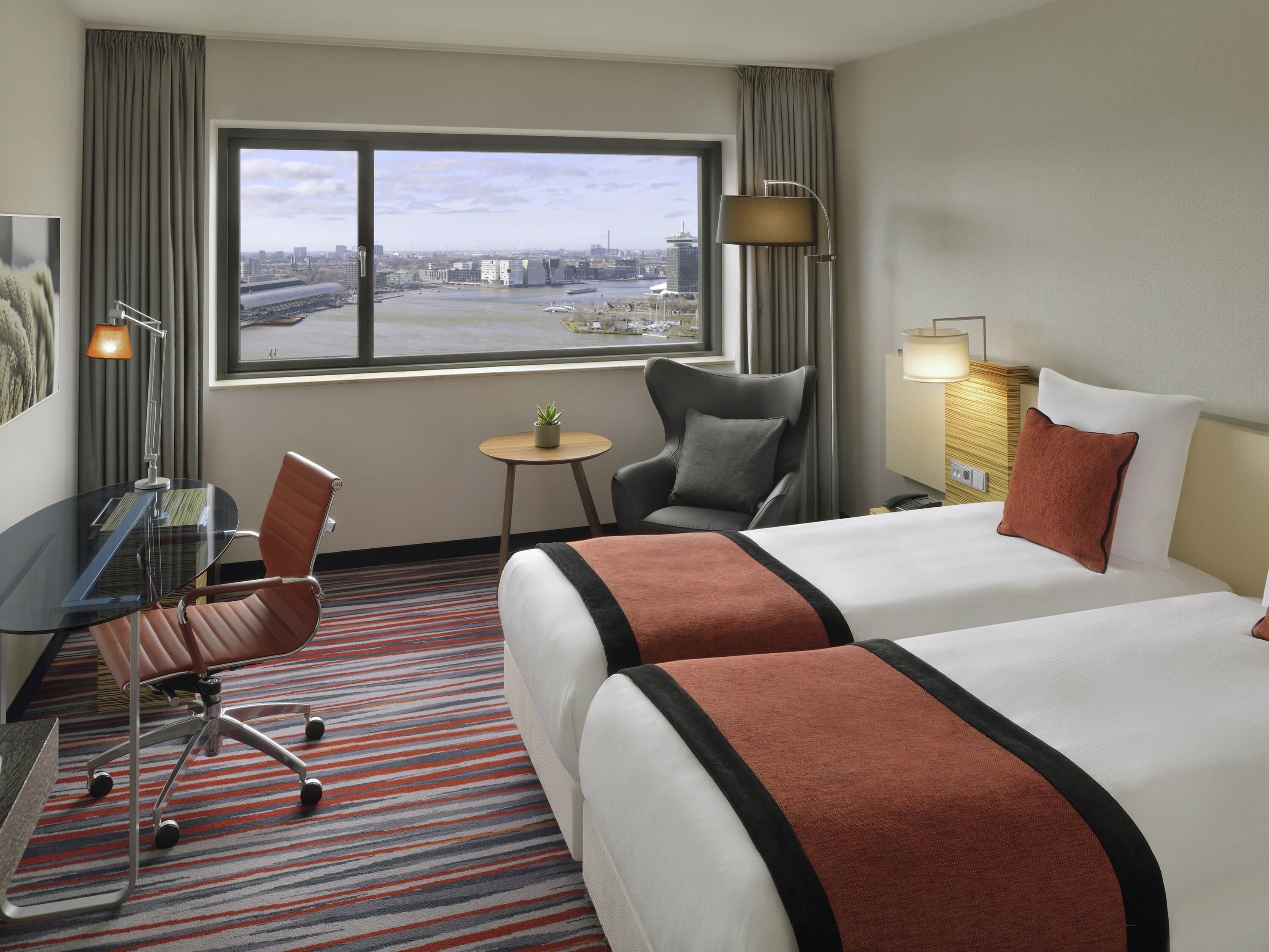Habitación de hotel con dos camas, un escritorio con silla, una mesita con planta y vistas a un paisaje urbano a través de un ventanal grande.