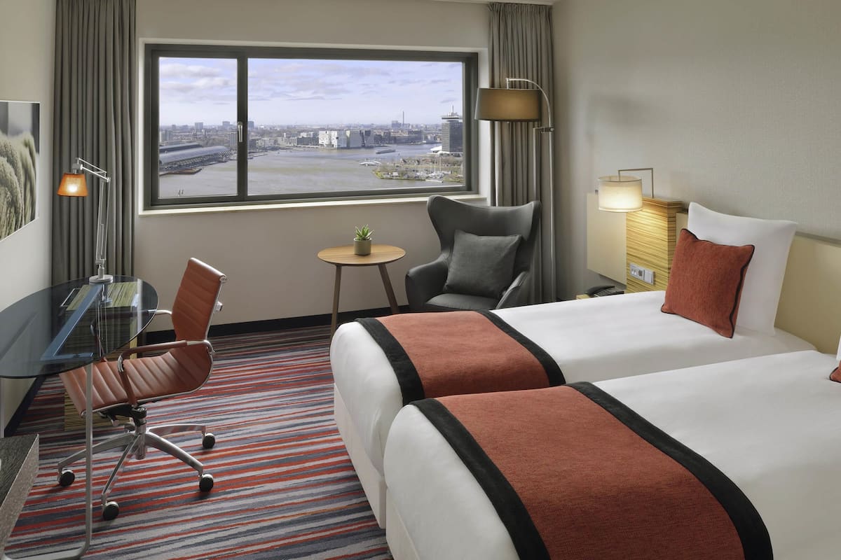 A hotel room with two beds, a desk with a chair, a small table with a plant, and a view of a cityscape through a large window.