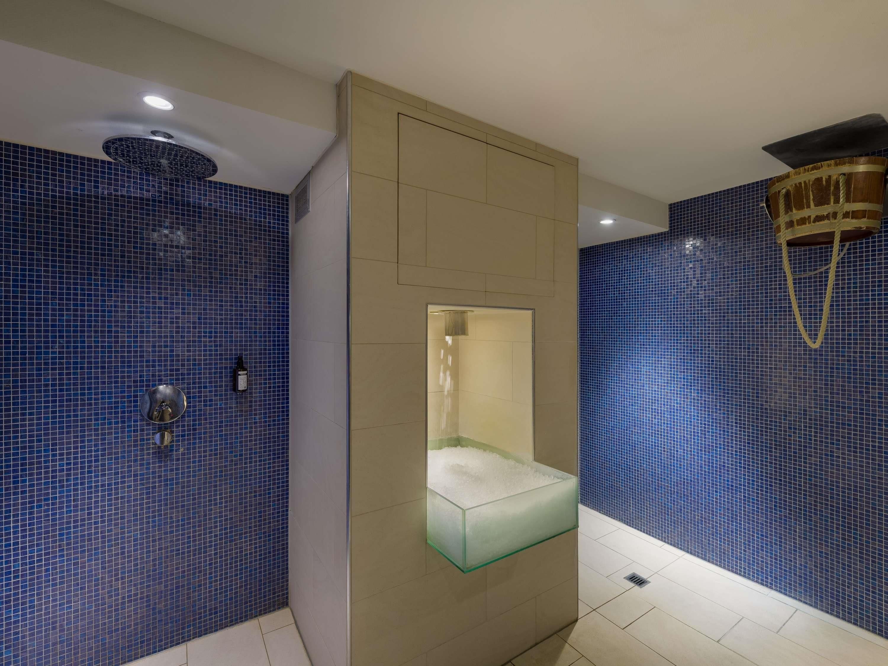 A bathroom with blue mosaic tiles, a glass shower enclosure, a beige wall, and a ceiling light fixture.
