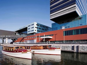 Un edificio moderno con fachada roja y azul, un barco amarrado frente a él y un puente al fondo.