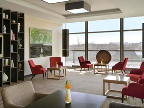A modern hotel lobby with red chairs, a large painting, and a view of a cityscape through large windows.