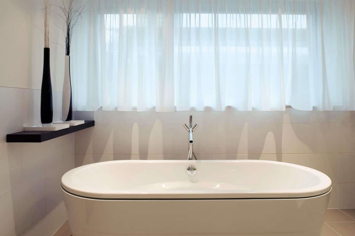 A modern bathroom with a freestanding bathtub, a showerhead, and a window with sheer curtains.