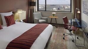 A hotel room with a large bed, a desk, a chair, a small table with flowers, and a view of a cityscape through a large window. - Mövenpick Hotel Amsterdam City Centre (Amsterdam)