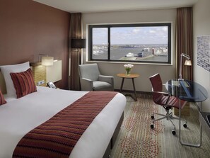 A hotel room with a large bed, a desk, a chair, a small table with flowers, and a view of a cityscape through a large window. - Mövenpick Hotel Amsterdam City Centre (Amsterdam)