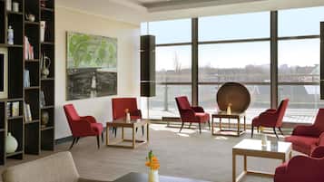 Een moderne hotellobby met rode stoelen, een groot schilderij en uitzicht op een stadsgezicht door grote ramen.