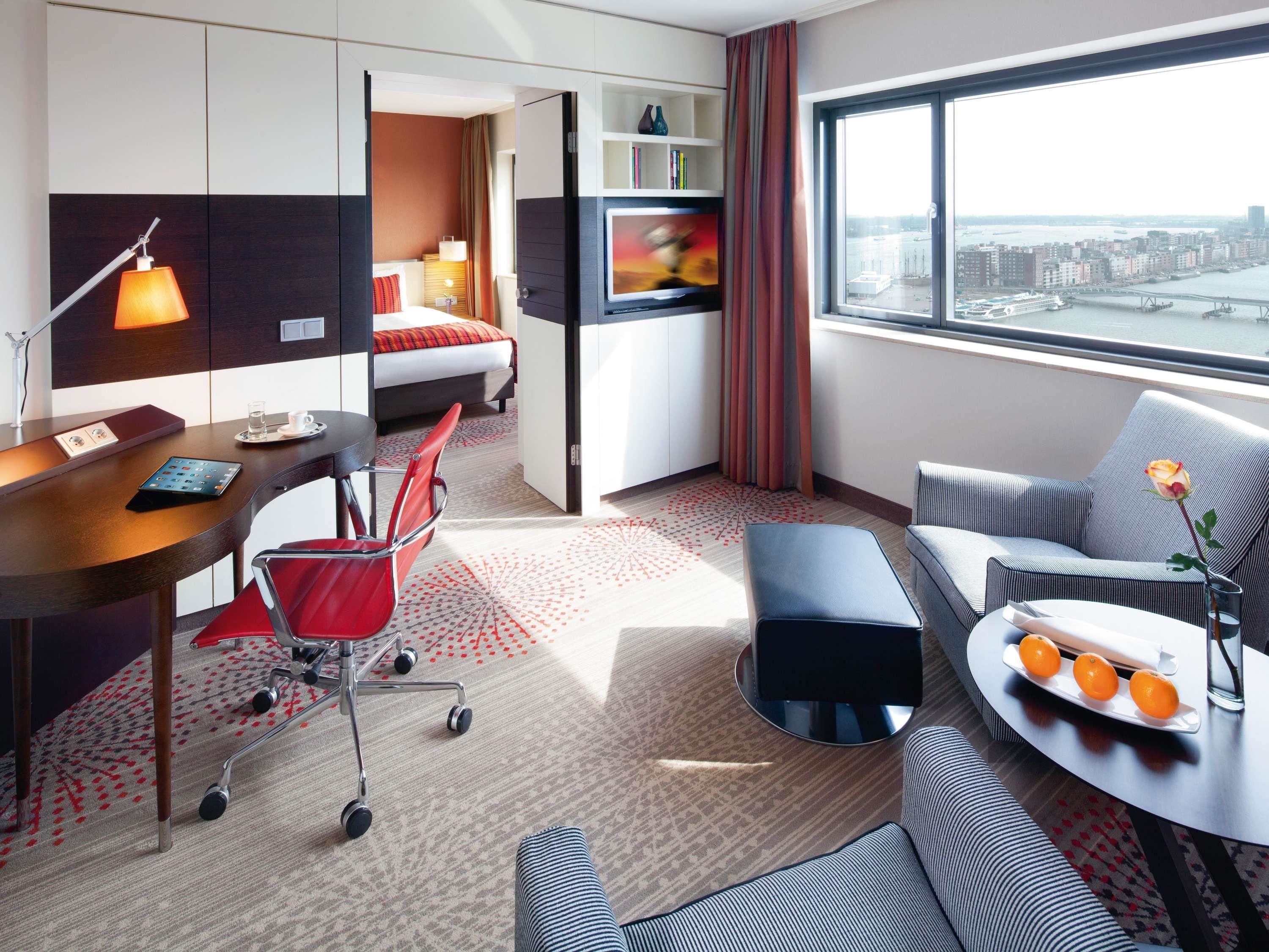 A hotel room with a bed, desk, chair, sofa, and a view of the city.