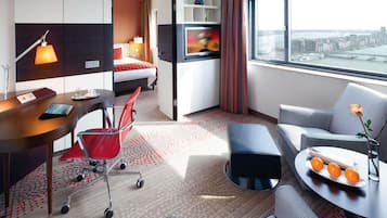 A hotel room with a bed, desk, chair, sofa, and a view of the city.