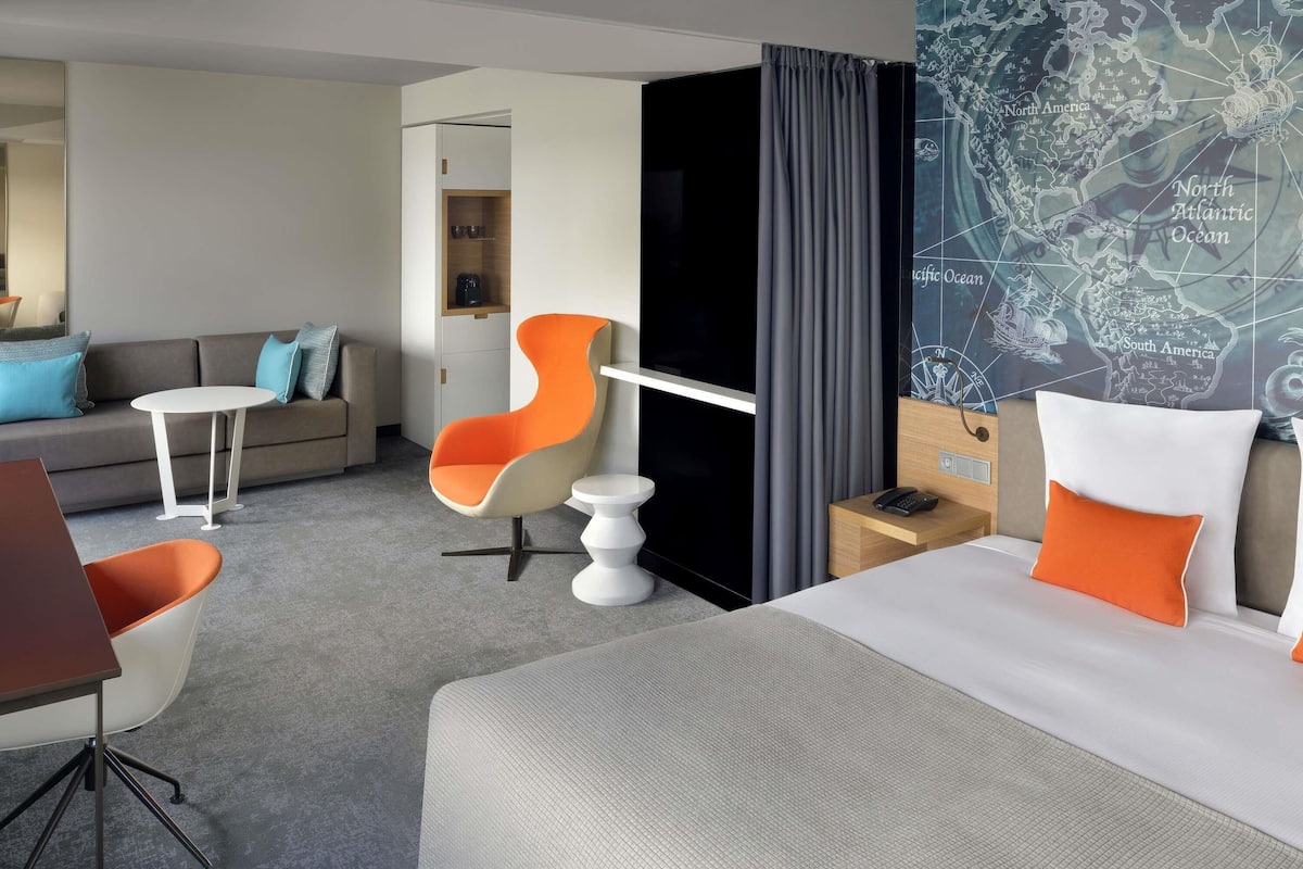 A modern hotel room with a bed, a desk, a chair, a small table, and a map on the wall.