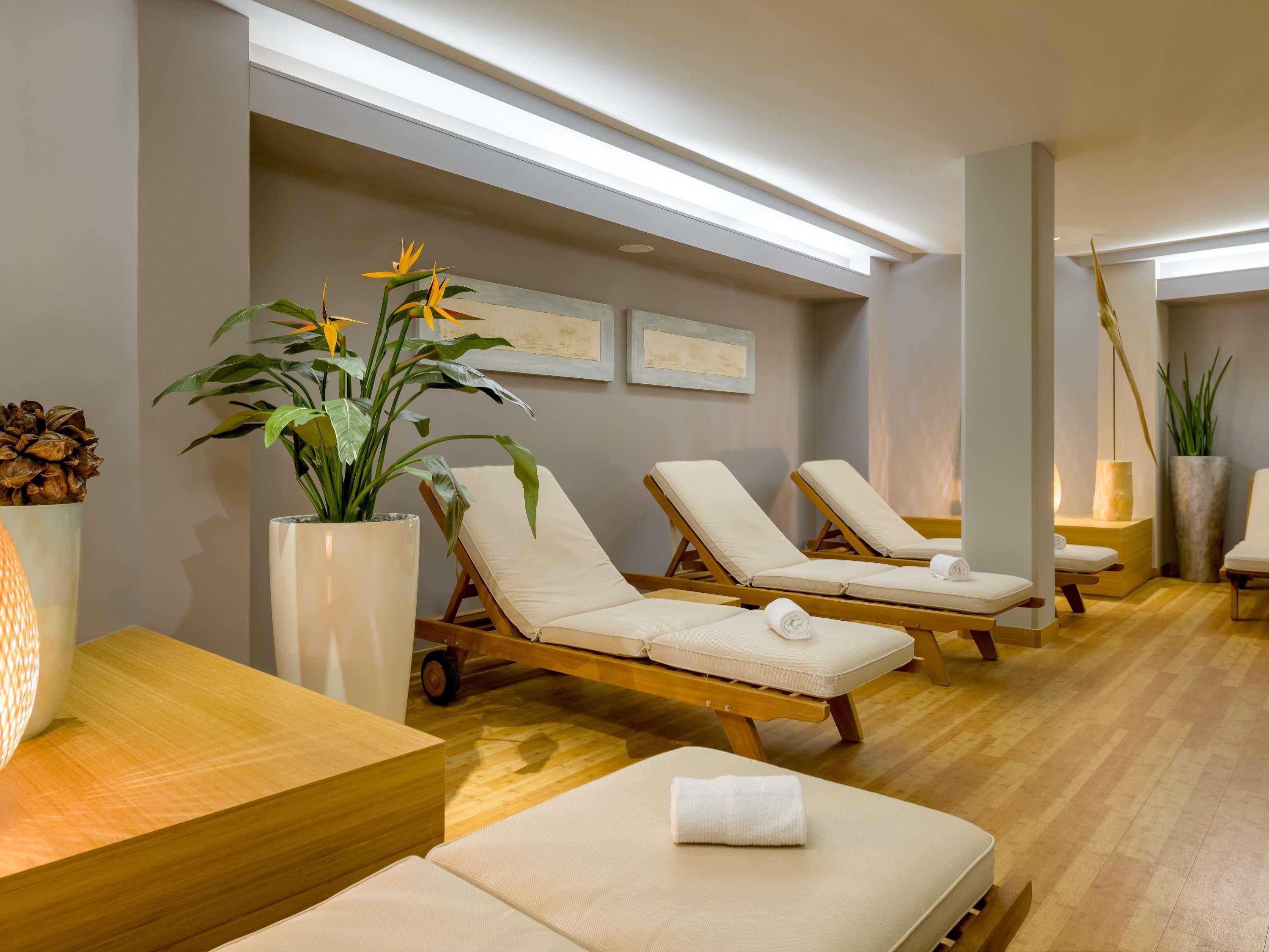 Ein Spa-Raum mit Holzboden, weißen gepolsterten Lounge-Stühlen, einer großen Topfpflanze und einem Holztisch.