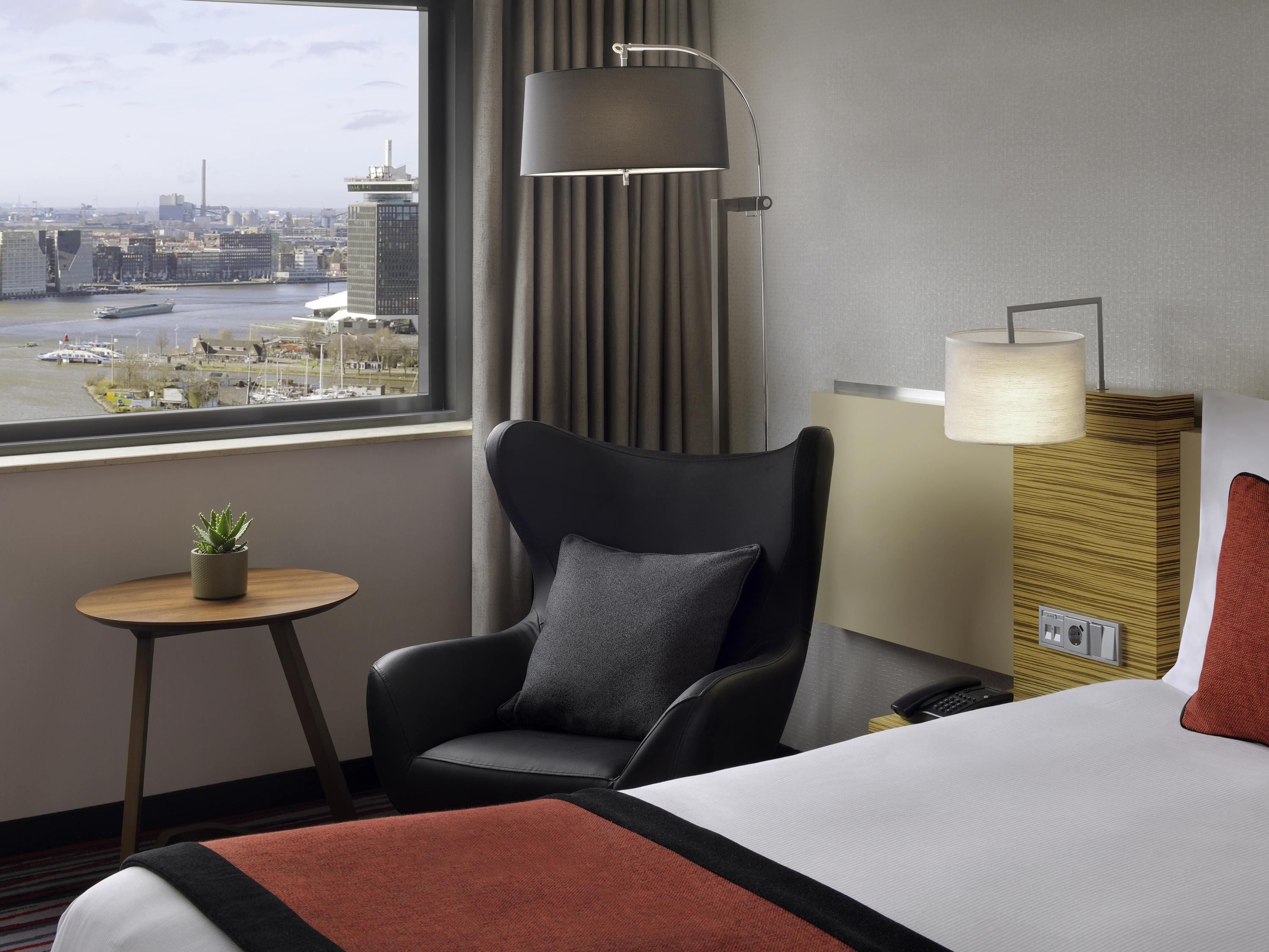 Habitación de hotel con cama, una silla, una mesita con una planta, una lámpara y vistas de la ciudad a través de una ventana.