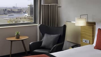 Hotelkamer met een bed, een stoel, een kleintje tafeltje met een plant, een lamp en uitzicht op een stadsbeeld door een raam.