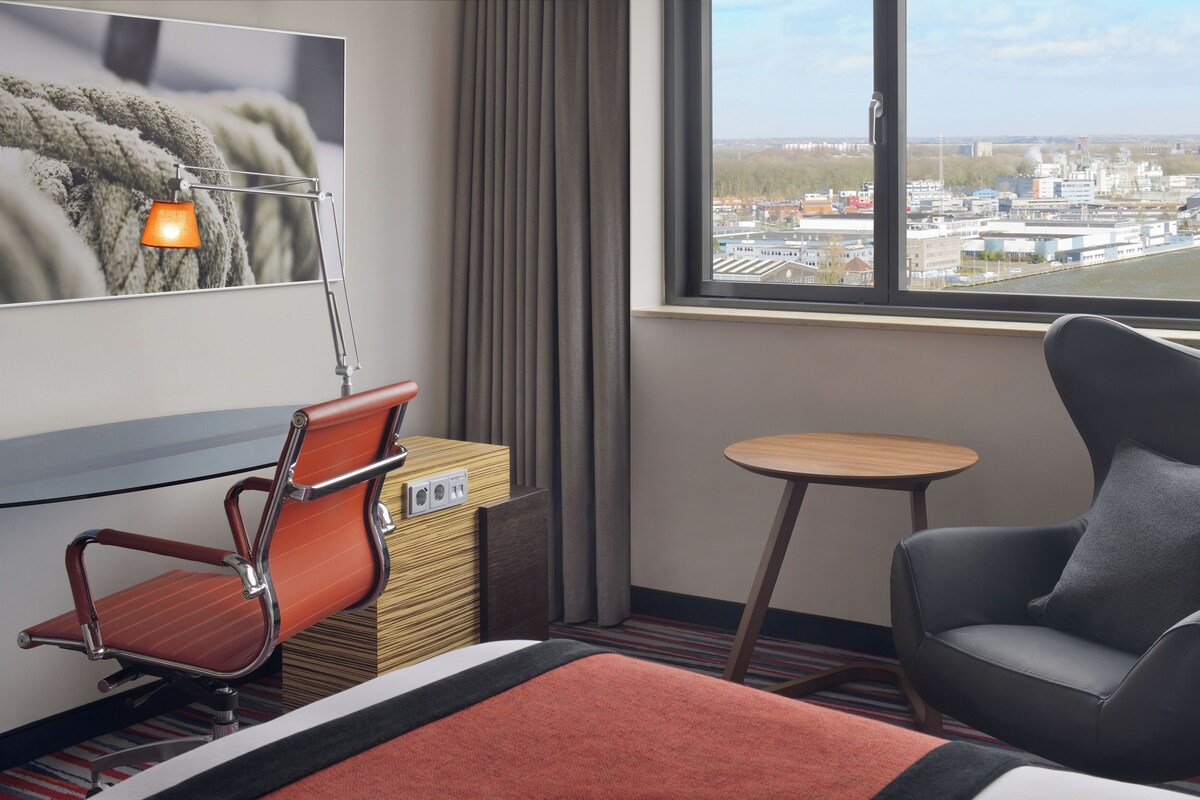 Habitación de hotel con cama, escritorio con silla, mesita, sofá, televisión y ventana con vista a la ciudad.