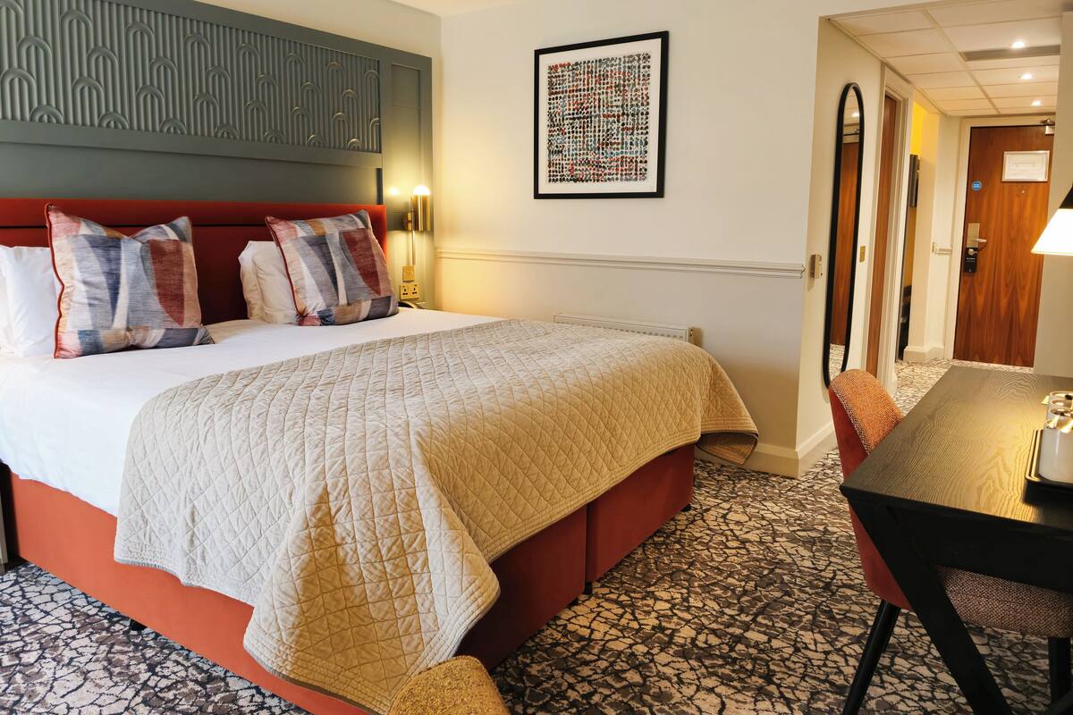 A hotel room with a large bed, a red upholstered headboard, a patterned rug, and a dining area with a table and chairs.