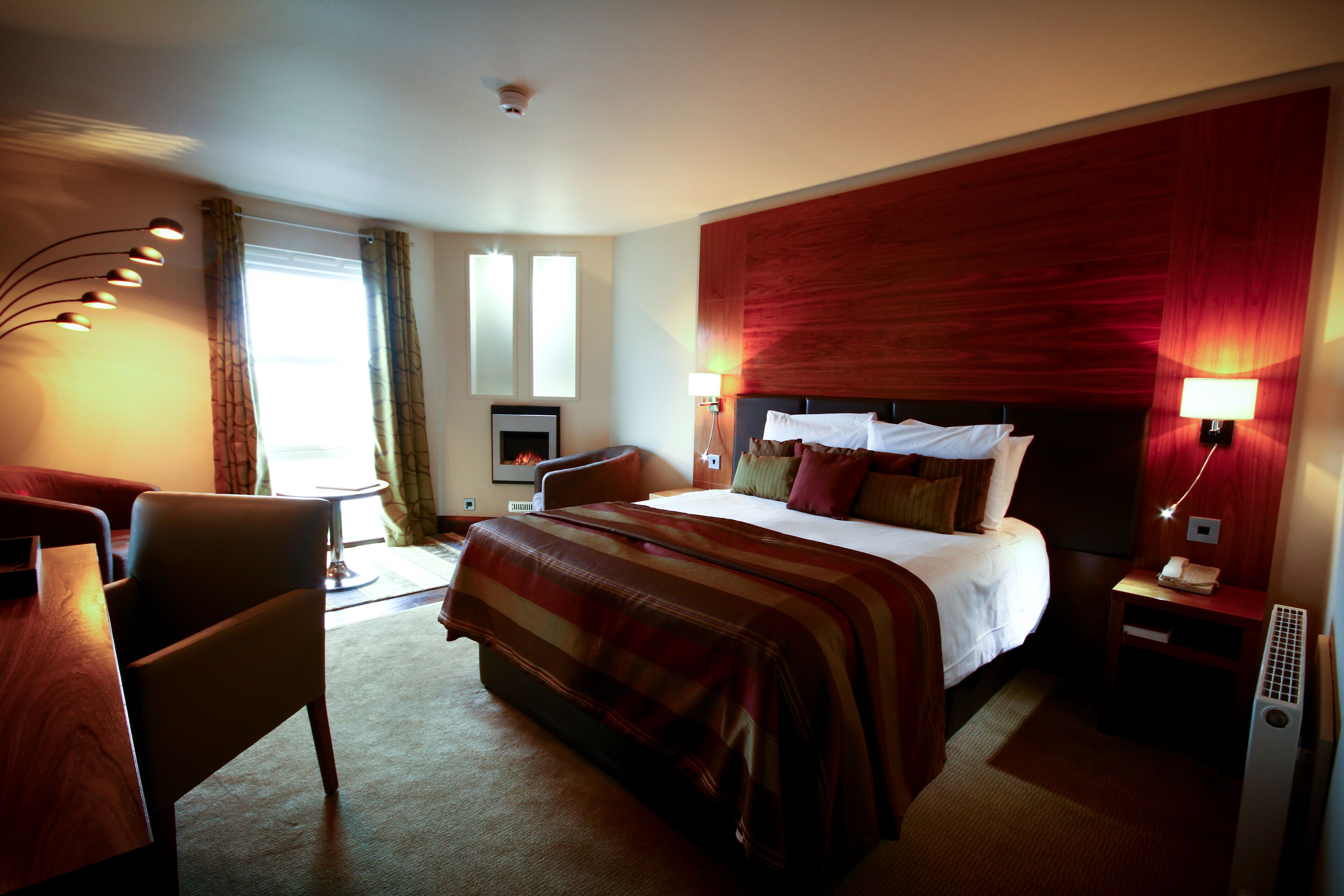 A hotel room with a large bed, a desk, a chair, a lamp, and a heater.