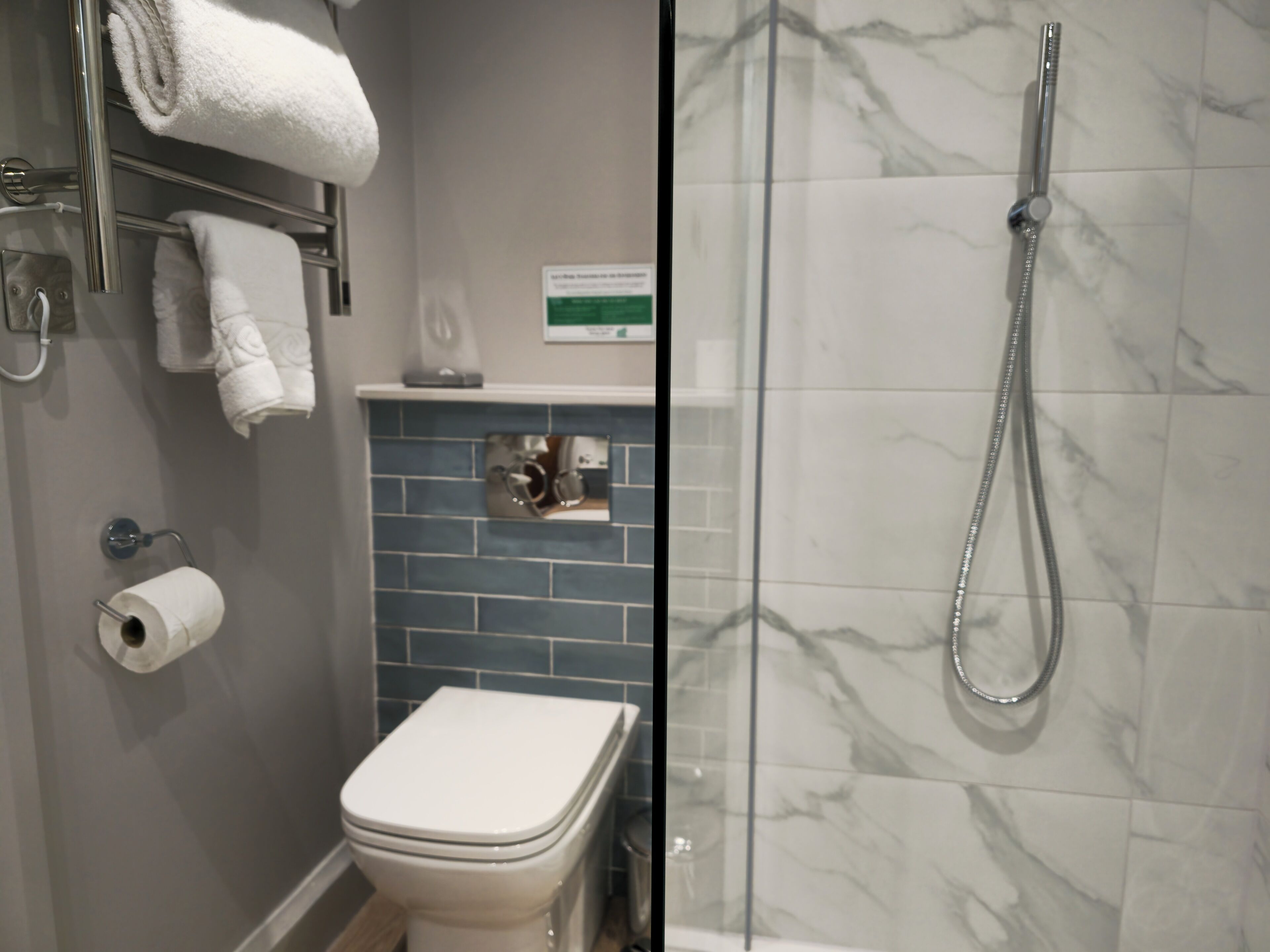 A bathroom with a toilet, a towel rack with towels, a paper towel dispenser, a shower with a glass door, and a sign on the wall.