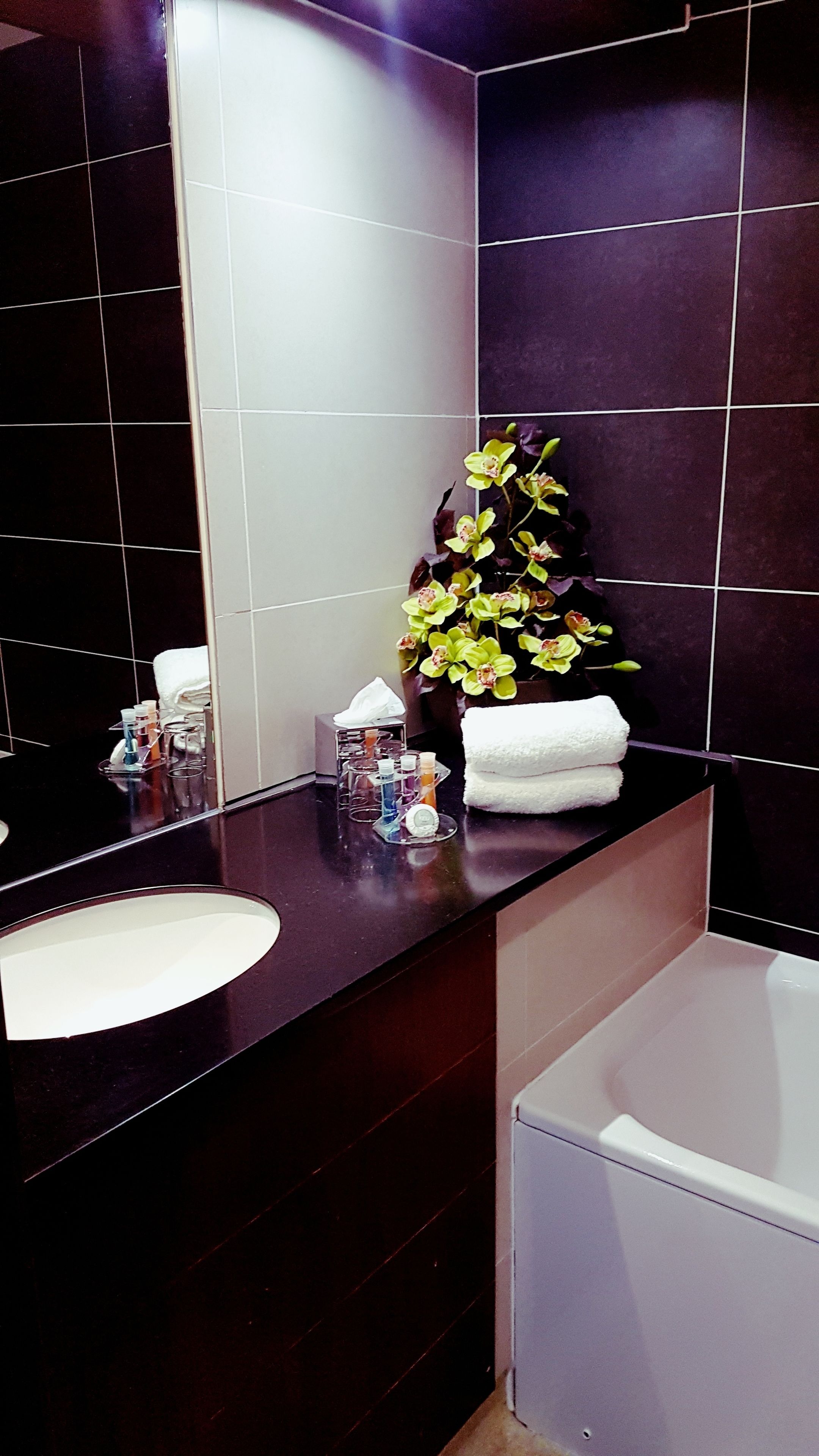 A modern bathroom with a dark countertop, a white sink, and a large mirror. There is a vase with flowers and a towel rack with towels.