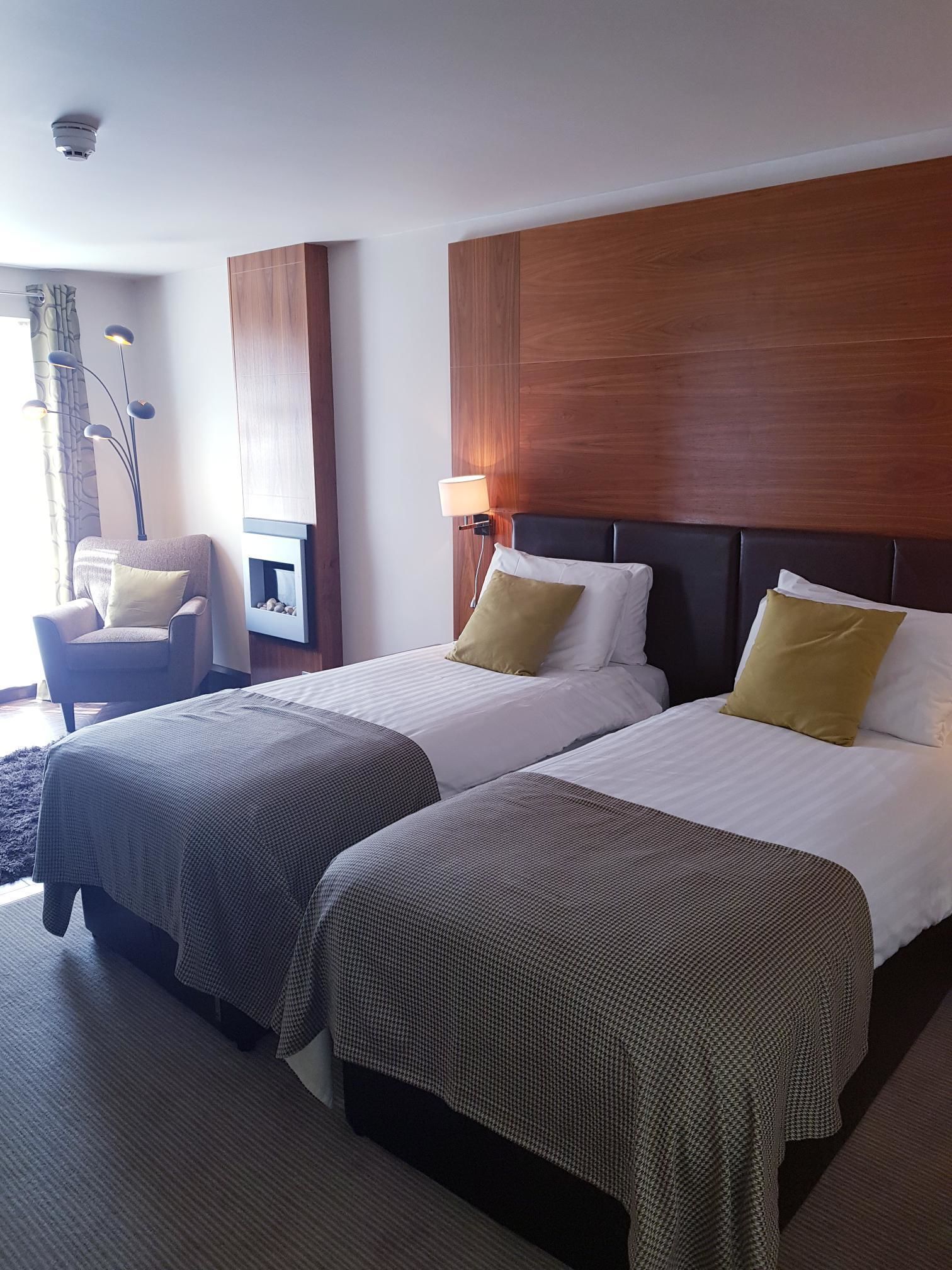 A hotel room with two beds, a chair, a lamp, and a wall-mounted TV.