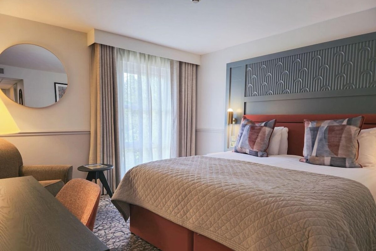 A hotel room with a large bed, a round mirror, a desk, and a chair.