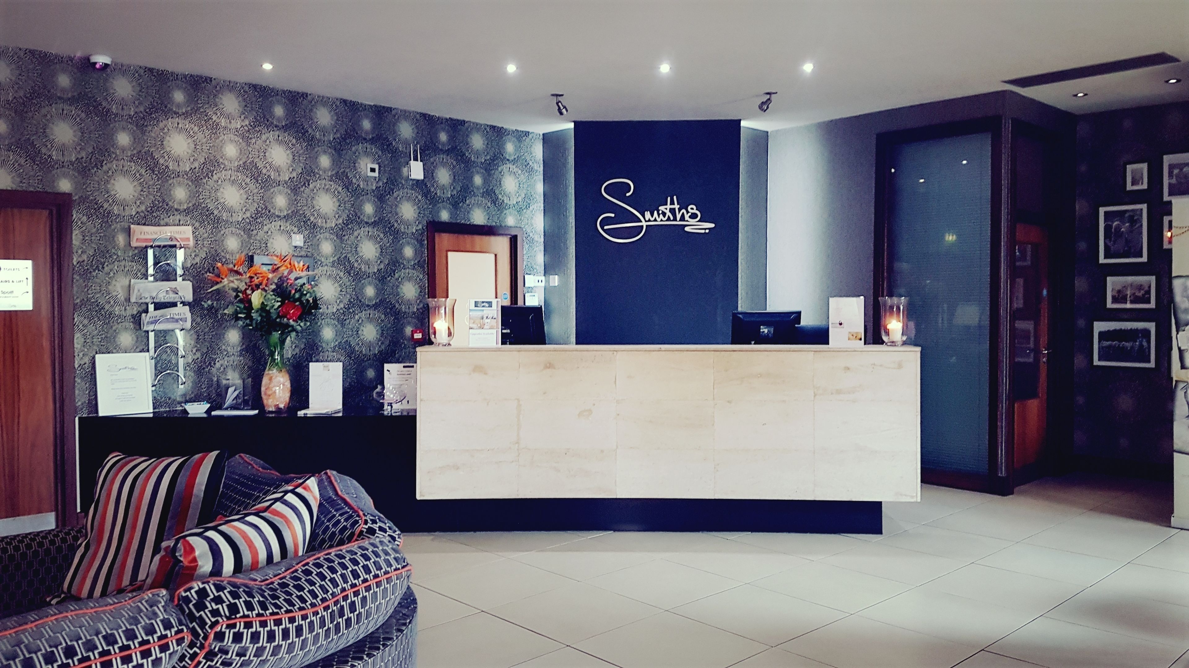 Un'area reception dal design moderno, con una parete blu scuro con su scritto 'Smiths' e un banco reception chiaro. Dietro il banco si nota una parete decorata con un motivo e, in primo piano, un divano con cuscini a righe.