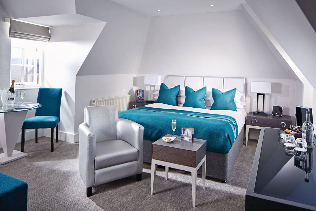 A modern hotel room with a bed, a desk, a chair, a small table, and a nightstand.
